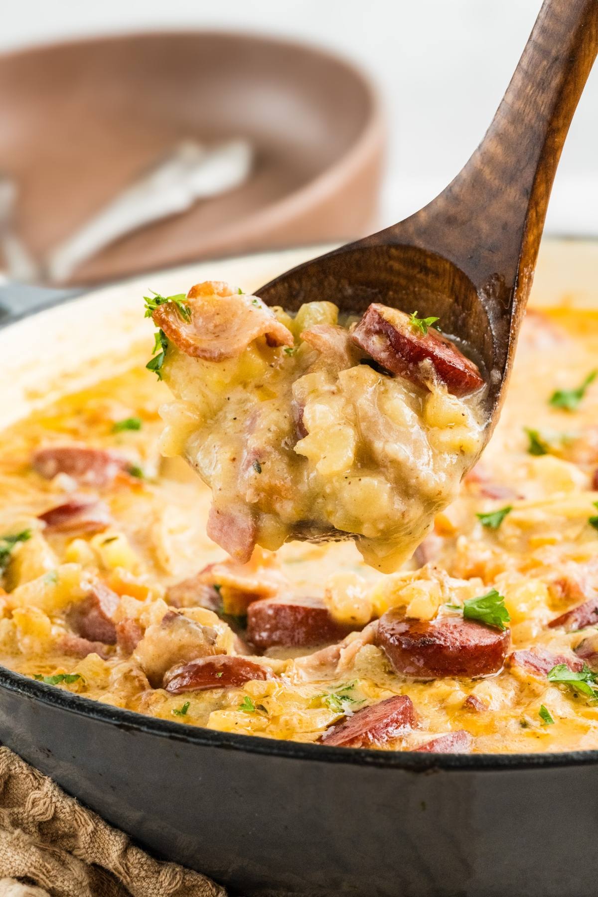 Wooden spoon scooping cheesy sausage and potato chowder from a casserole dish, garnished with parsley.