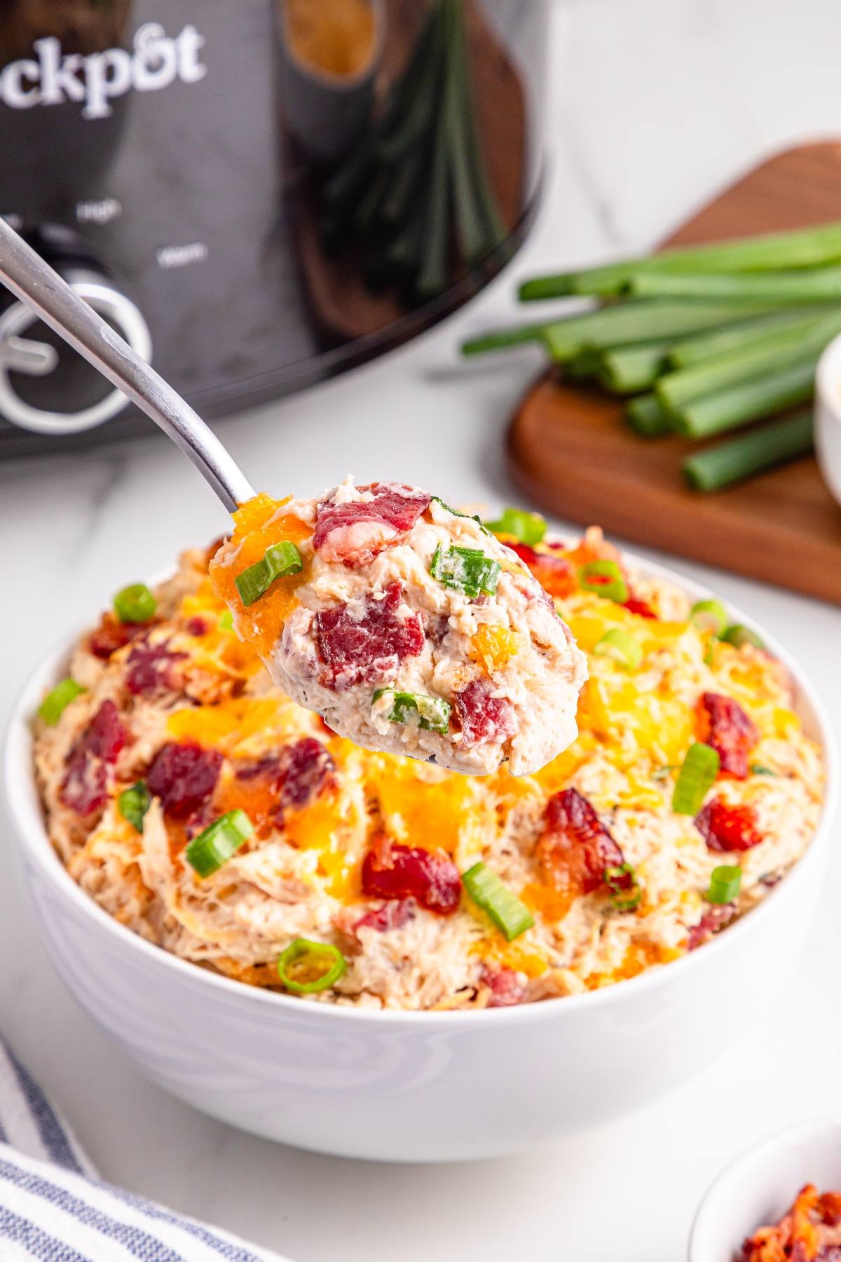 A spoonful of cheesy bacon ranch crack chicken lifted above a bowl, with green onions on top.