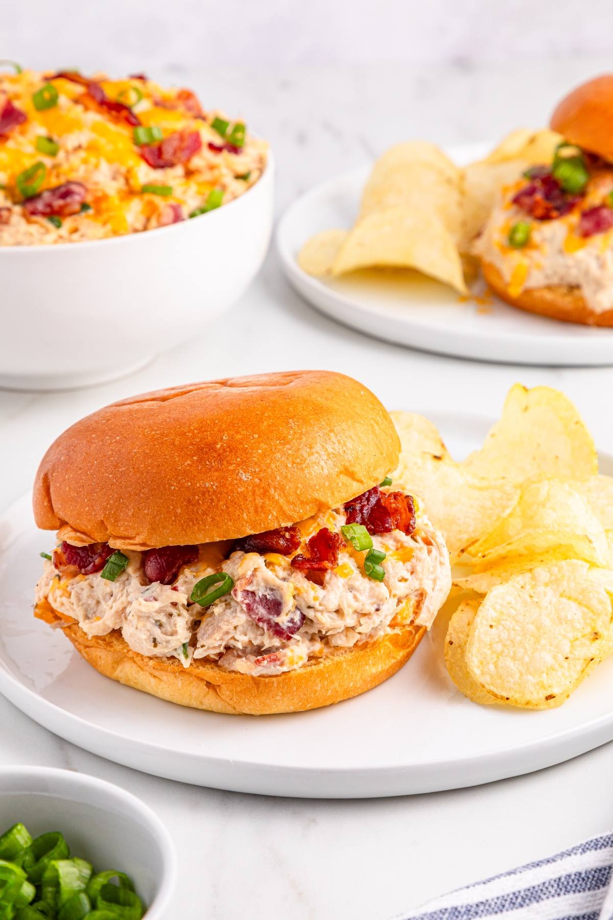 A creamy chicken bacon ranch sandwich on a plate with potato chips, with more dip and chips in the background.