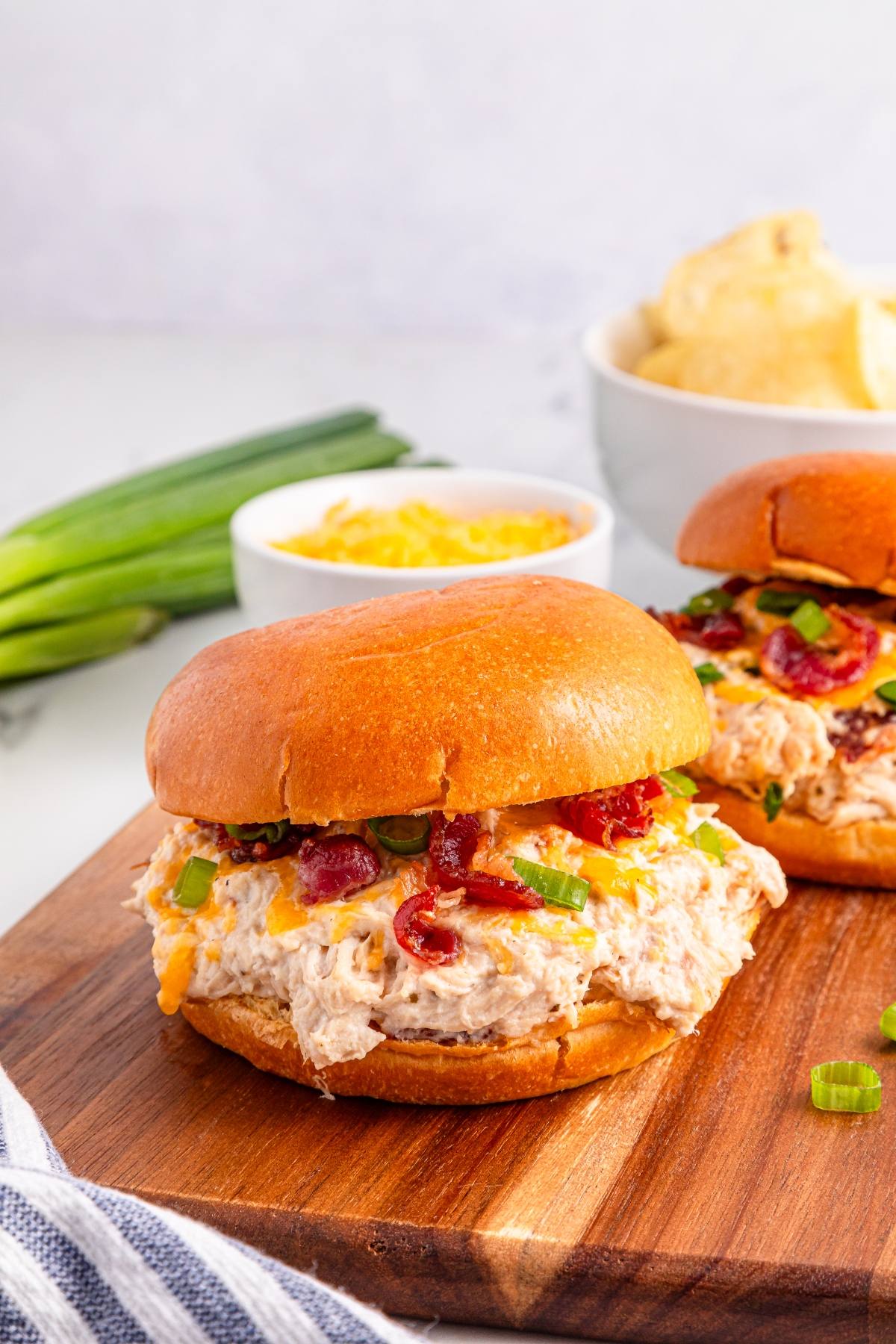 Chicken crack sandwiches with bacon, cheese, and green onions on a wooden board, chips and dip in background.