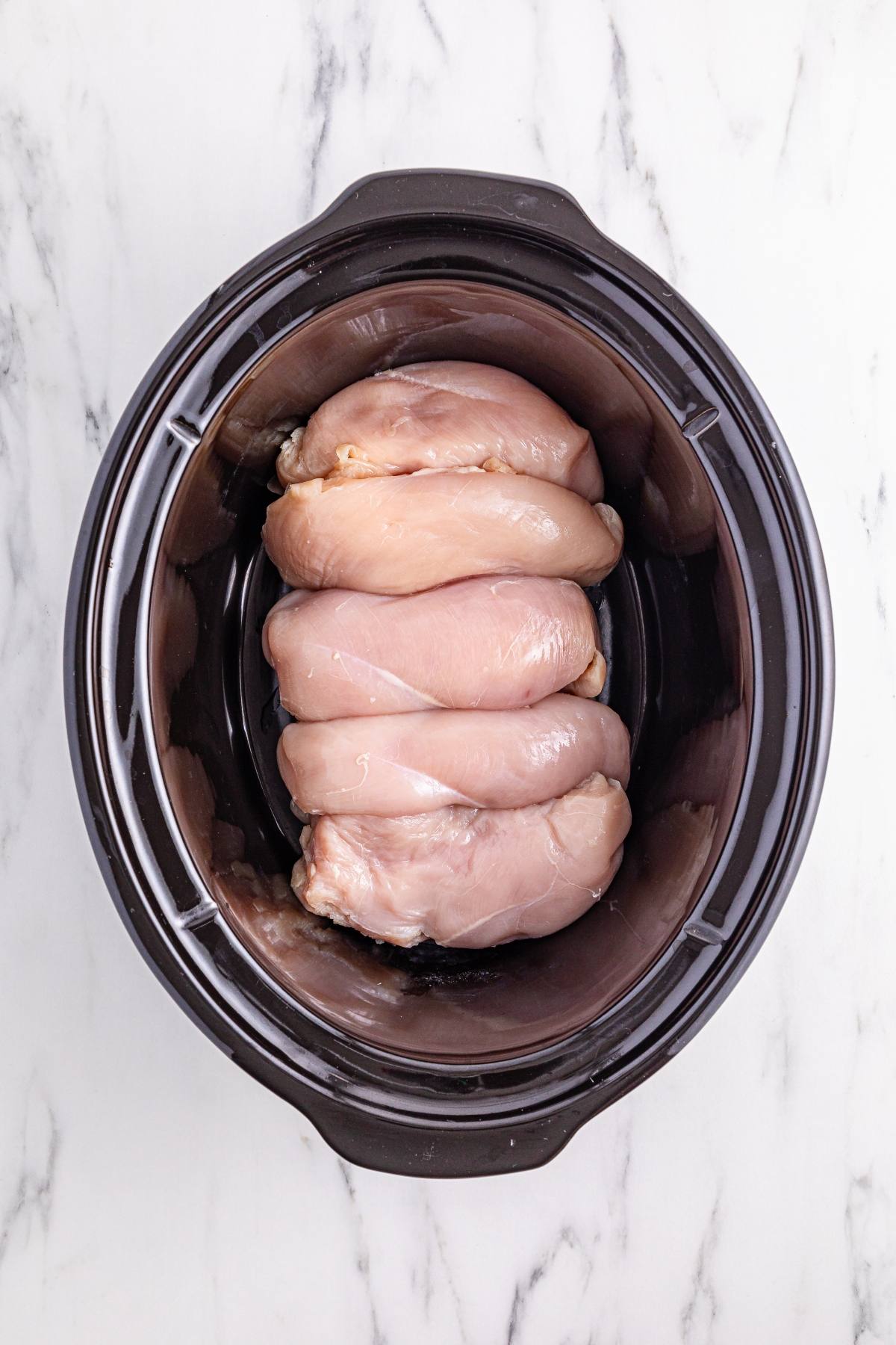 Raw chicken breasts stacked in a black slow cooker on a white marble surface.