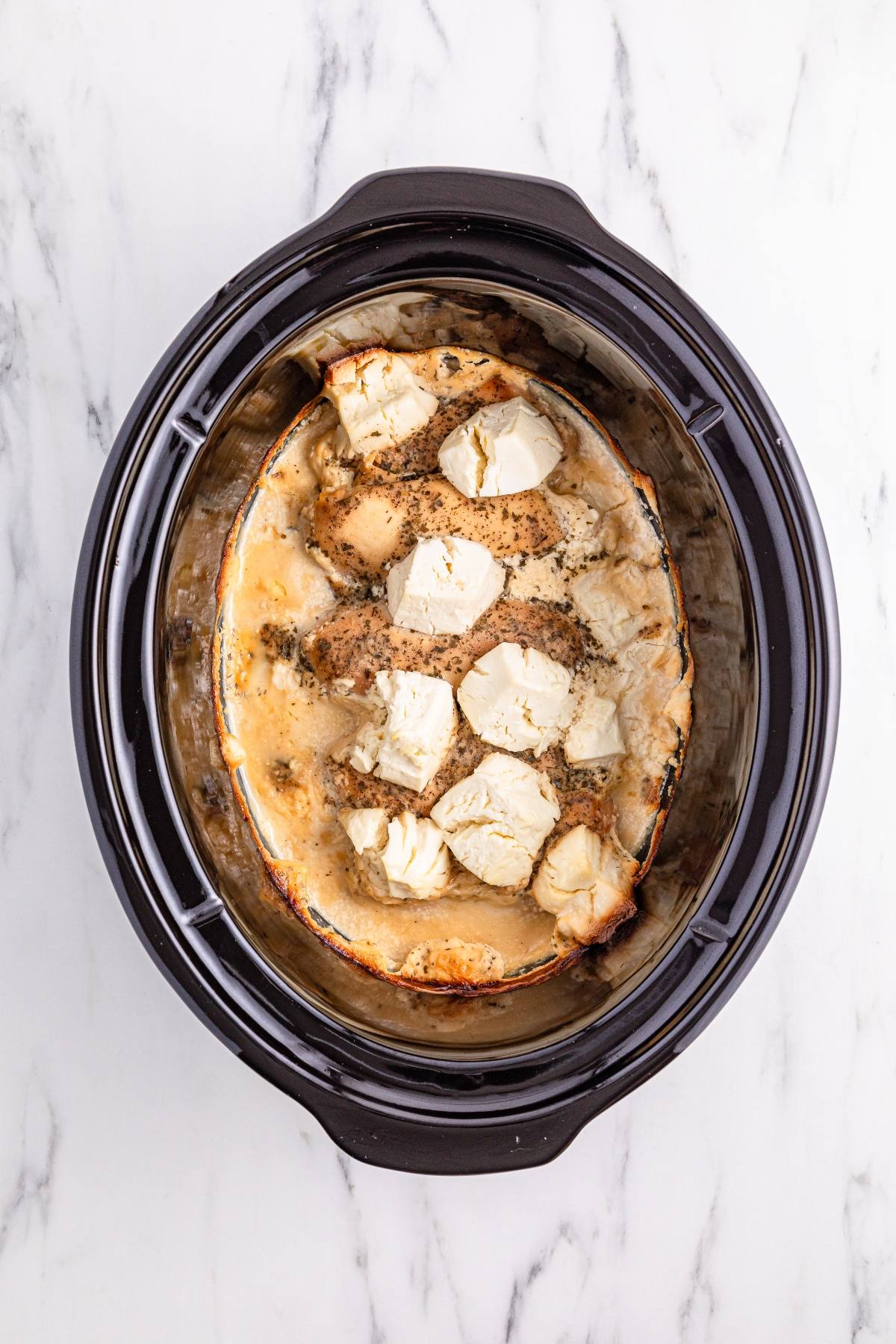 Chicken breasts with herbs and dollops of cream cheese cooking in a black slow cooker on a marble surface.