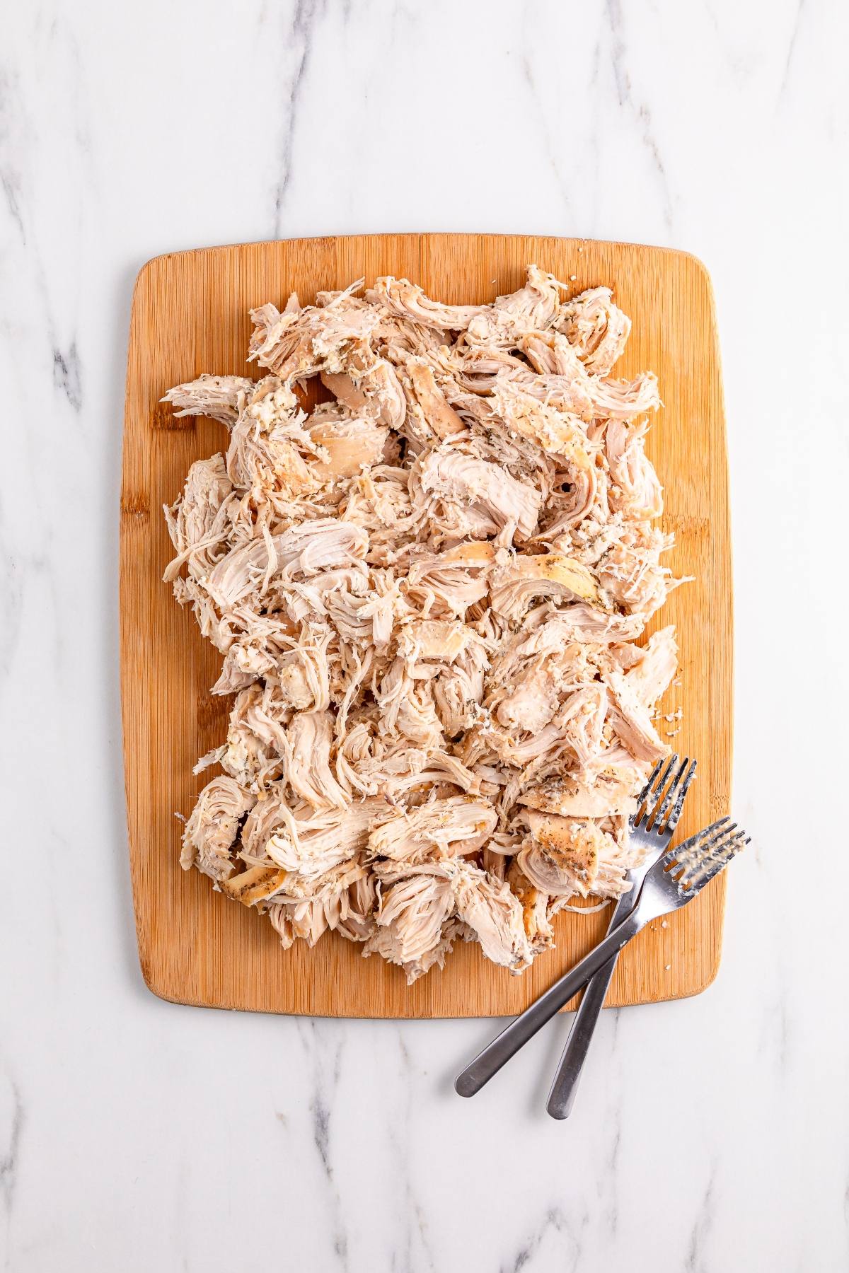 Shredded chicken piled on a wooden cutting board with two forks beside it on a marble surface.