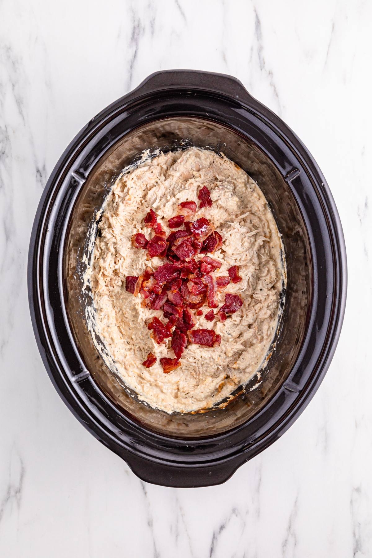 Shredded creamy mixture topped with chopped bacon in a black slow cooker on a marble surface.