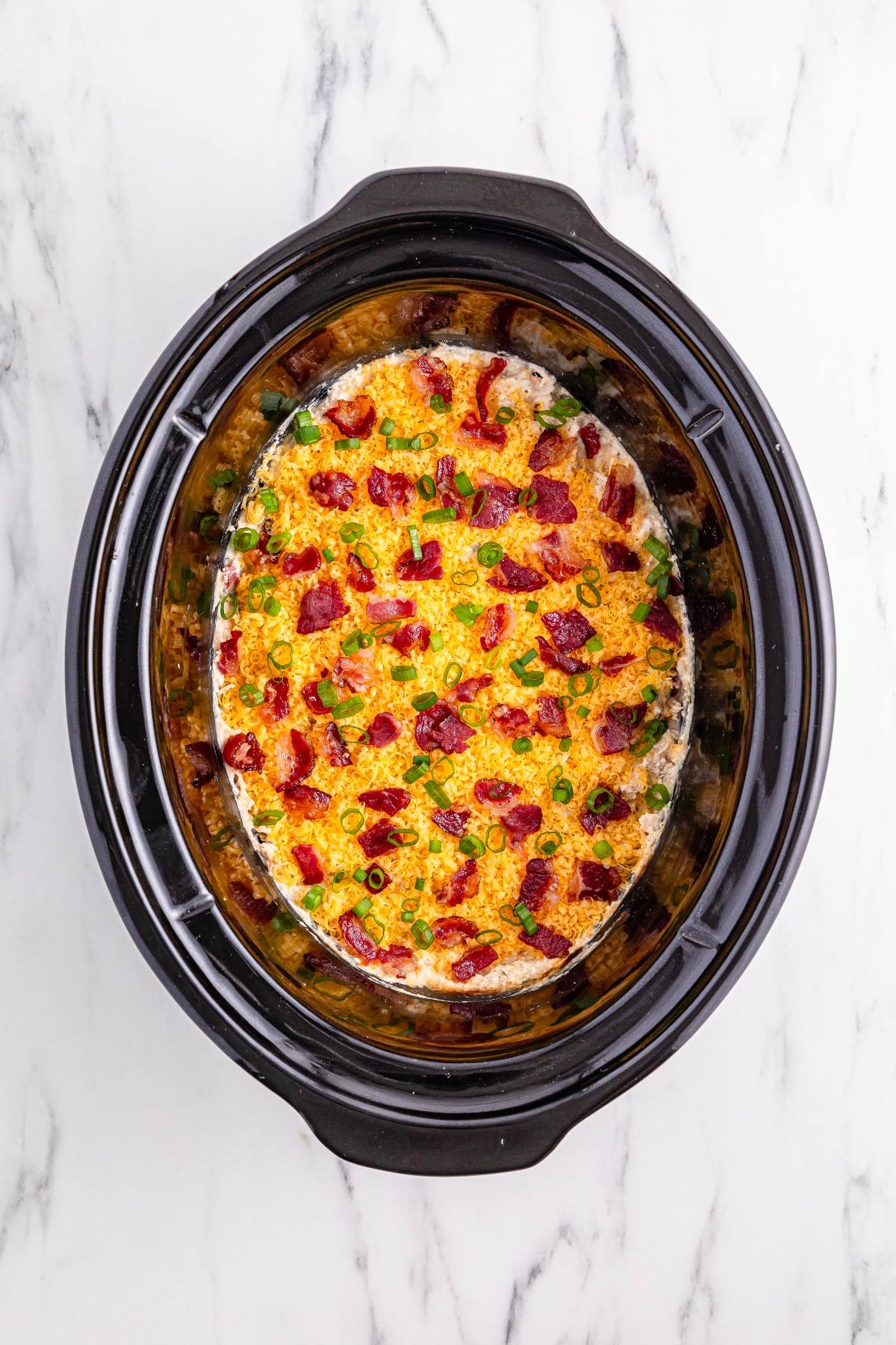 Casserole topped with bacon and green onions in a black slow cooker on a white marble surface.