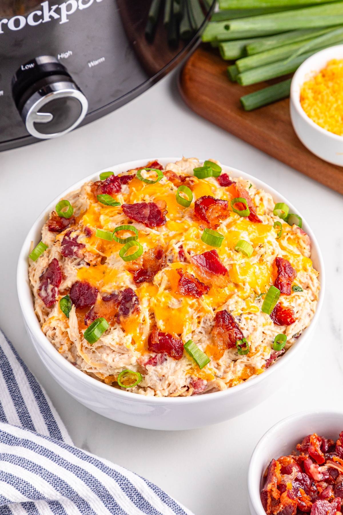 A bowl of creamy crack chicken with bacon, cheddar cheese, and green onions, next to a crockpot and fresh ingredients.