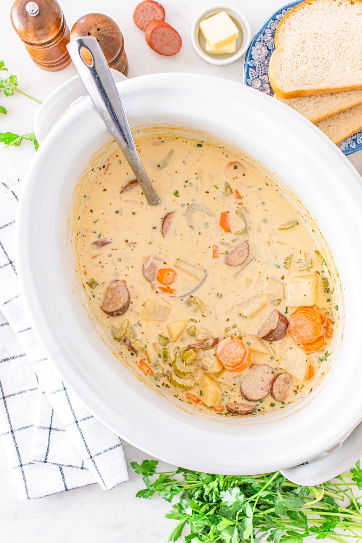 Creamy soup with sliced sausage, carrots, and potatoes in a white slow cooker with a serving spoon.