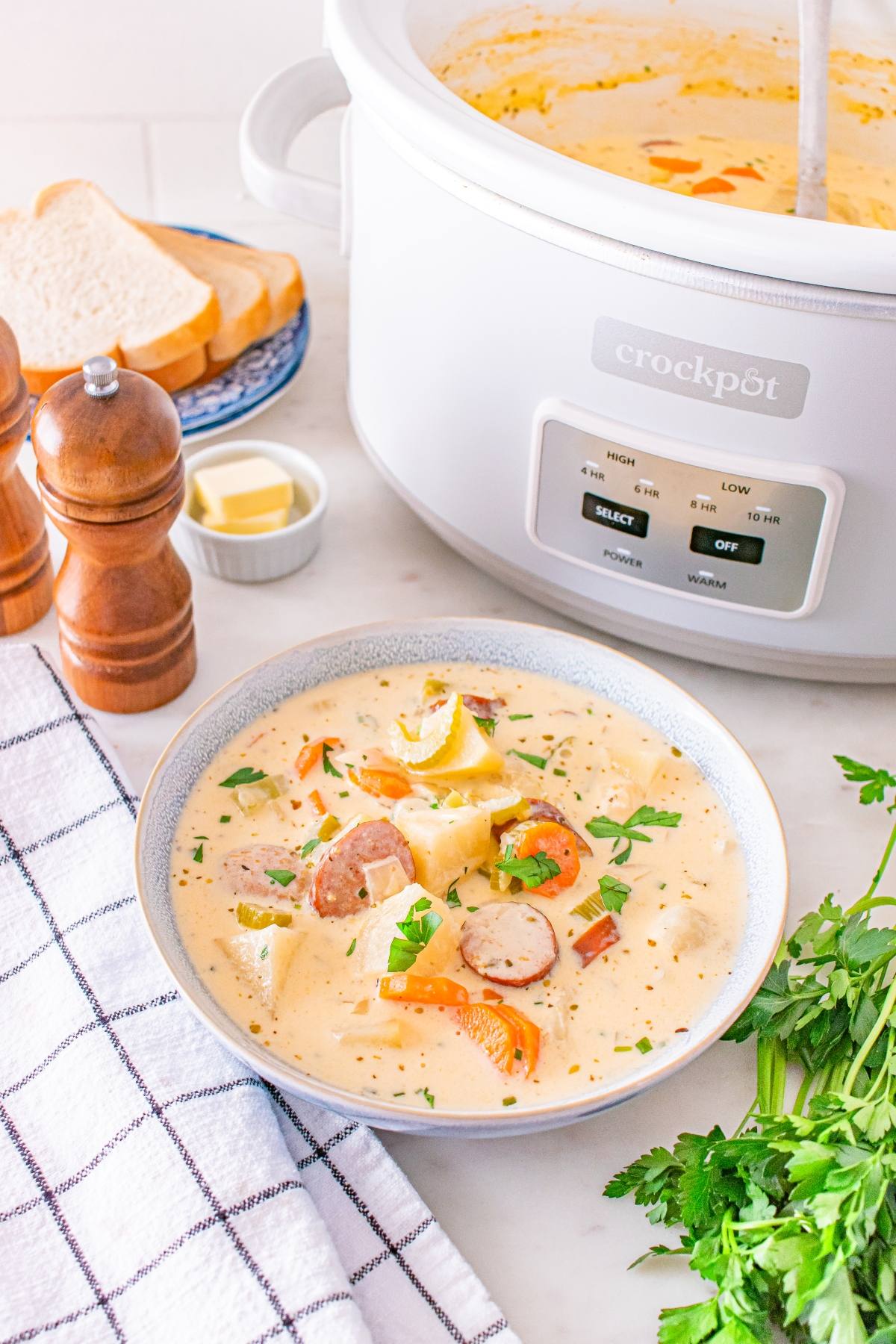 Bowl of creamy sausage and potato soup garnished with herbs, with a slow cooker, bread, and seasonings nearby.