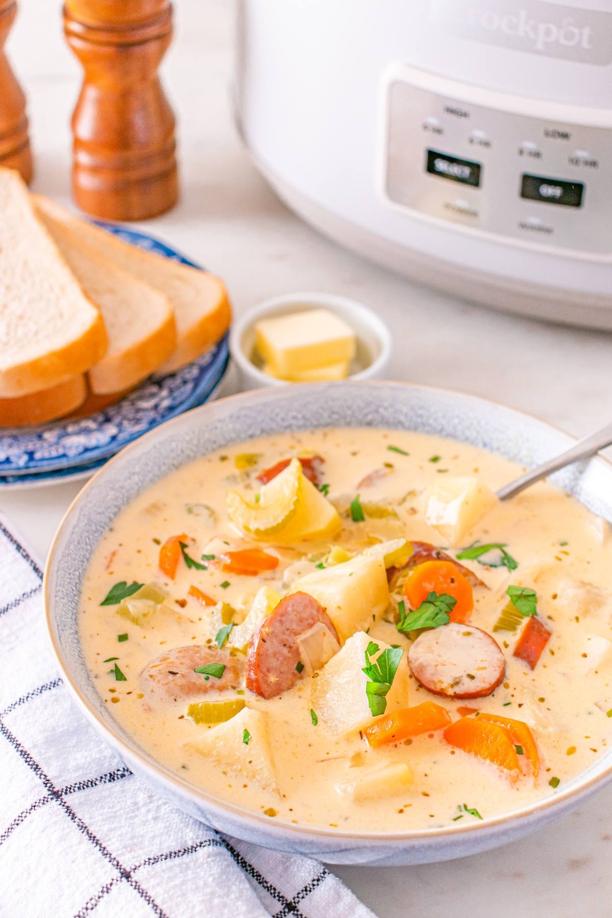 Creamy sausage and potato soup in a bowl with bread slices, butter, and a slow cooker in the background.