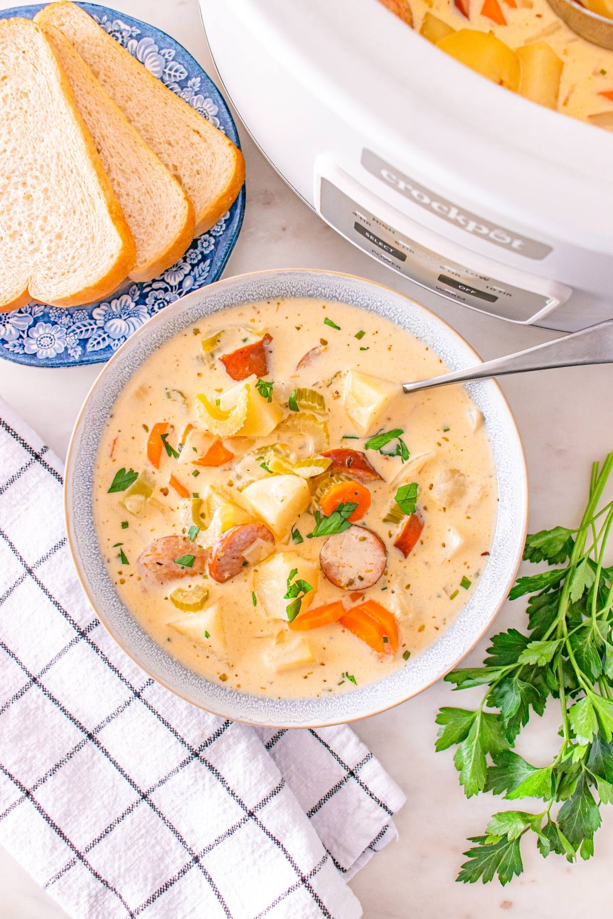 Bowl of creamy sausage and potato soup with a spoon, sliced bread, and fresh parsley on the side.