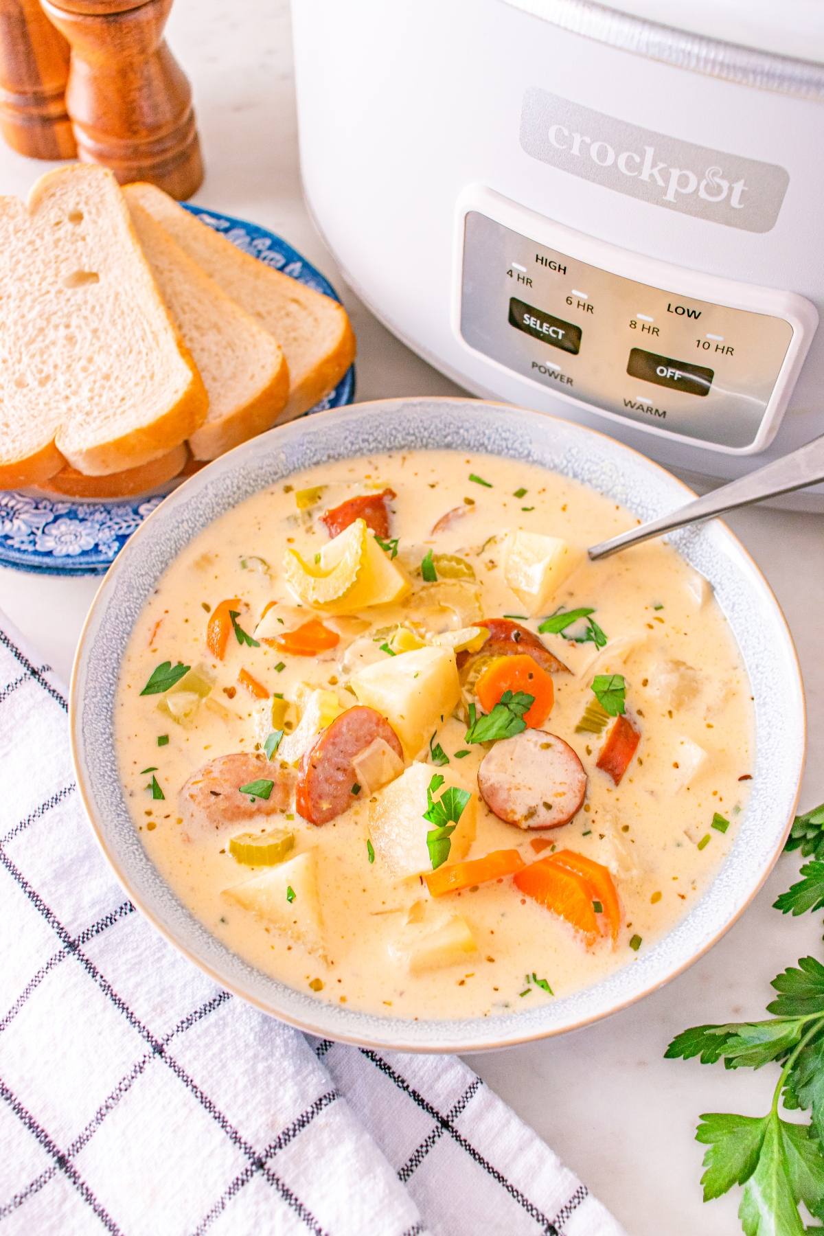 Bowl of creamy sausage and potato soup with herbs, next to sliced bread and a Crockpot on a checkered towel.