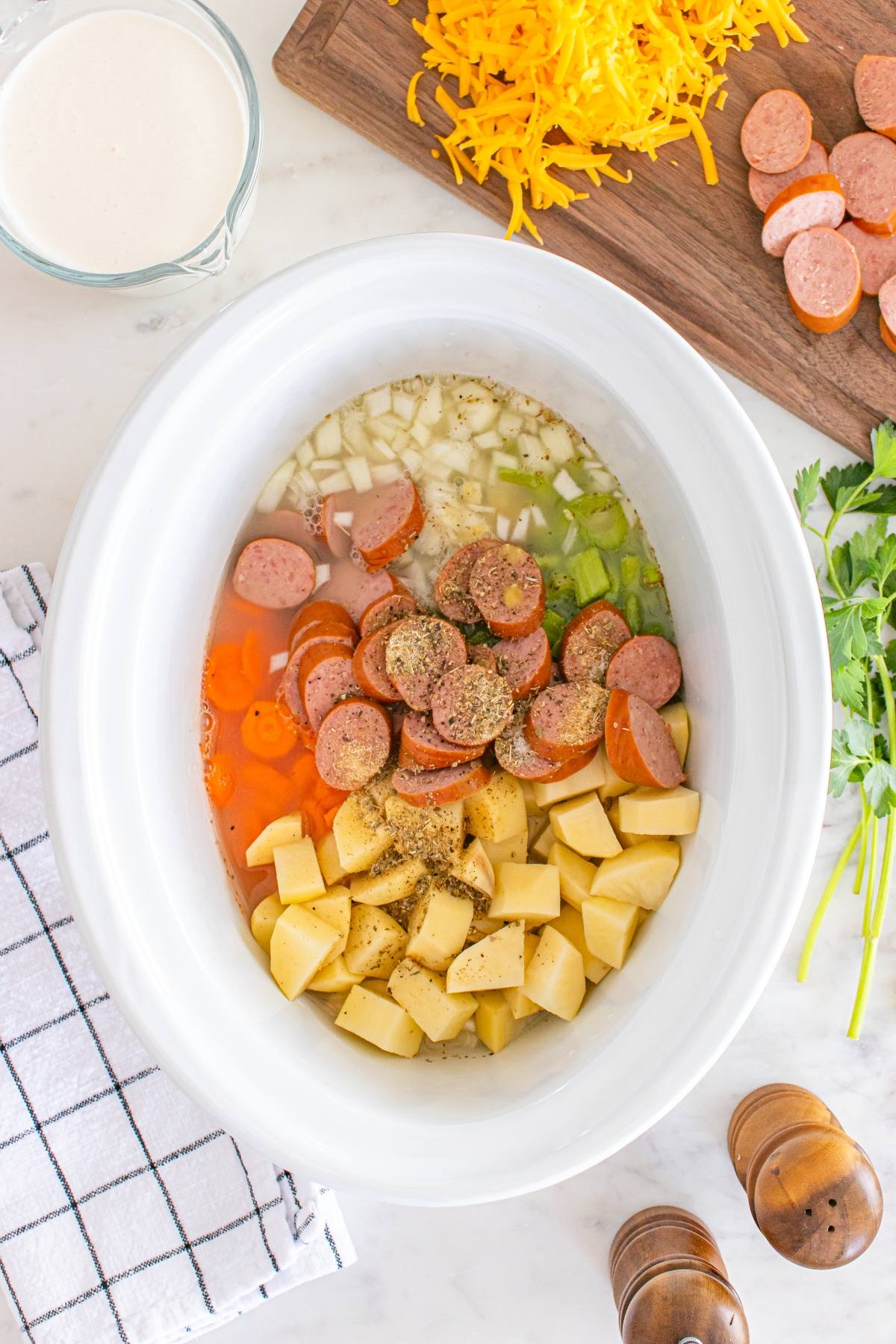 Slow cooker filled with potatoes, sausage, onions, celery, broth, and seasonings next to cheese and cutting board.