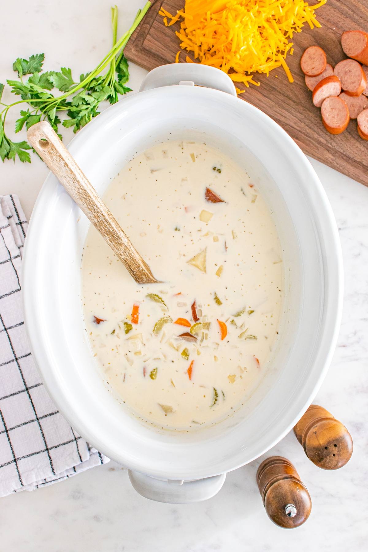 A crockpot filled with creamy soup, a wooden spoon, sliced sausage, shredded cheese, and parsley nearby.