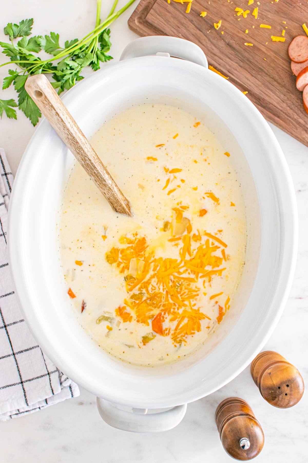 A creamy soup with shredded cheddar cheese in a white slow cooker, surrounded by herbs and a wooden spoon.