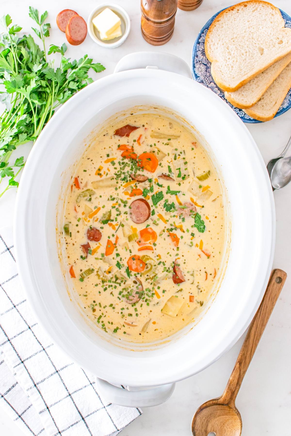 Creamy sausage and vegetable soup in a white slow cooker, surrounded by bread, parsley, and a wooden spoon.