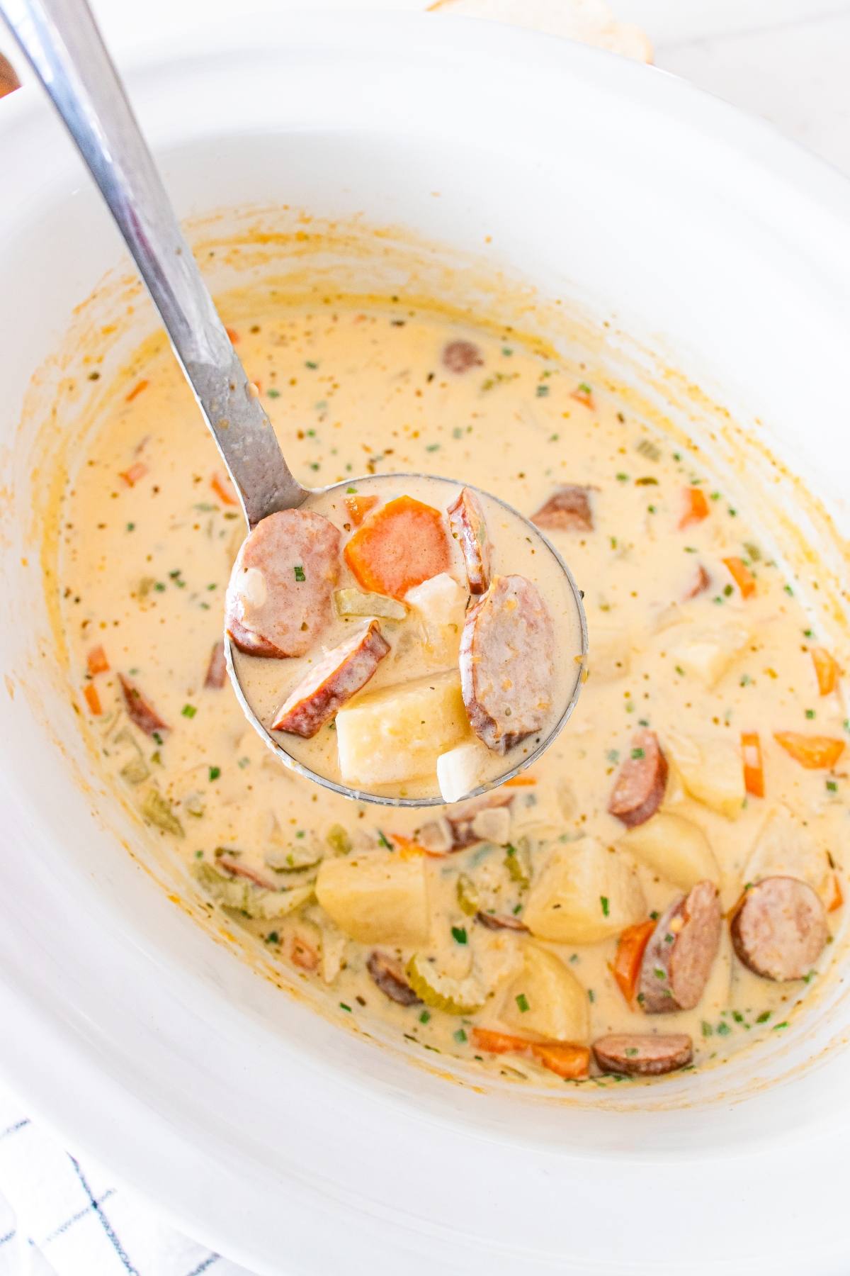 A ladle of creamy sausage and potato soup held over a white slow cooker.