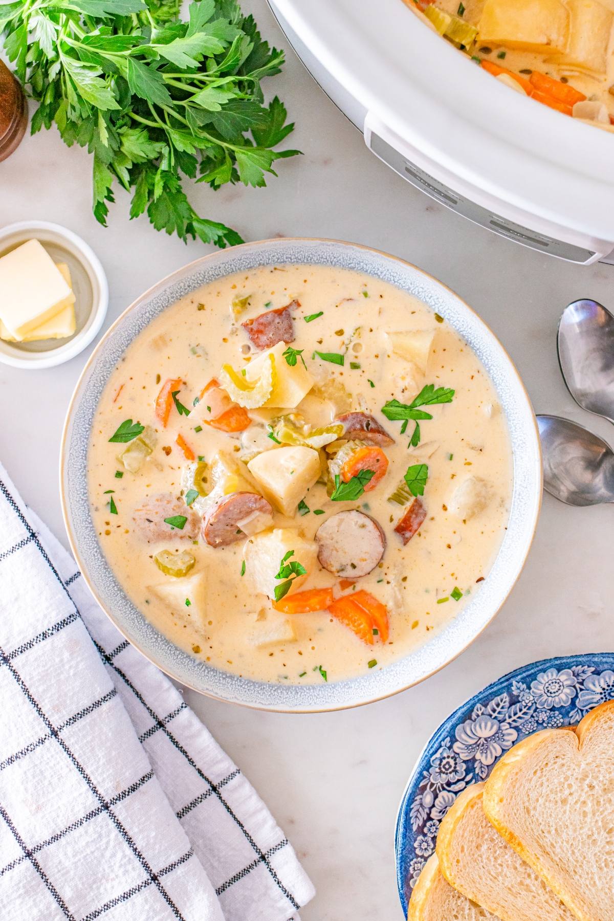 Creamy sausage and potato soup in a bowl, garnished with parsley, next to bread slices and fresh herbs.