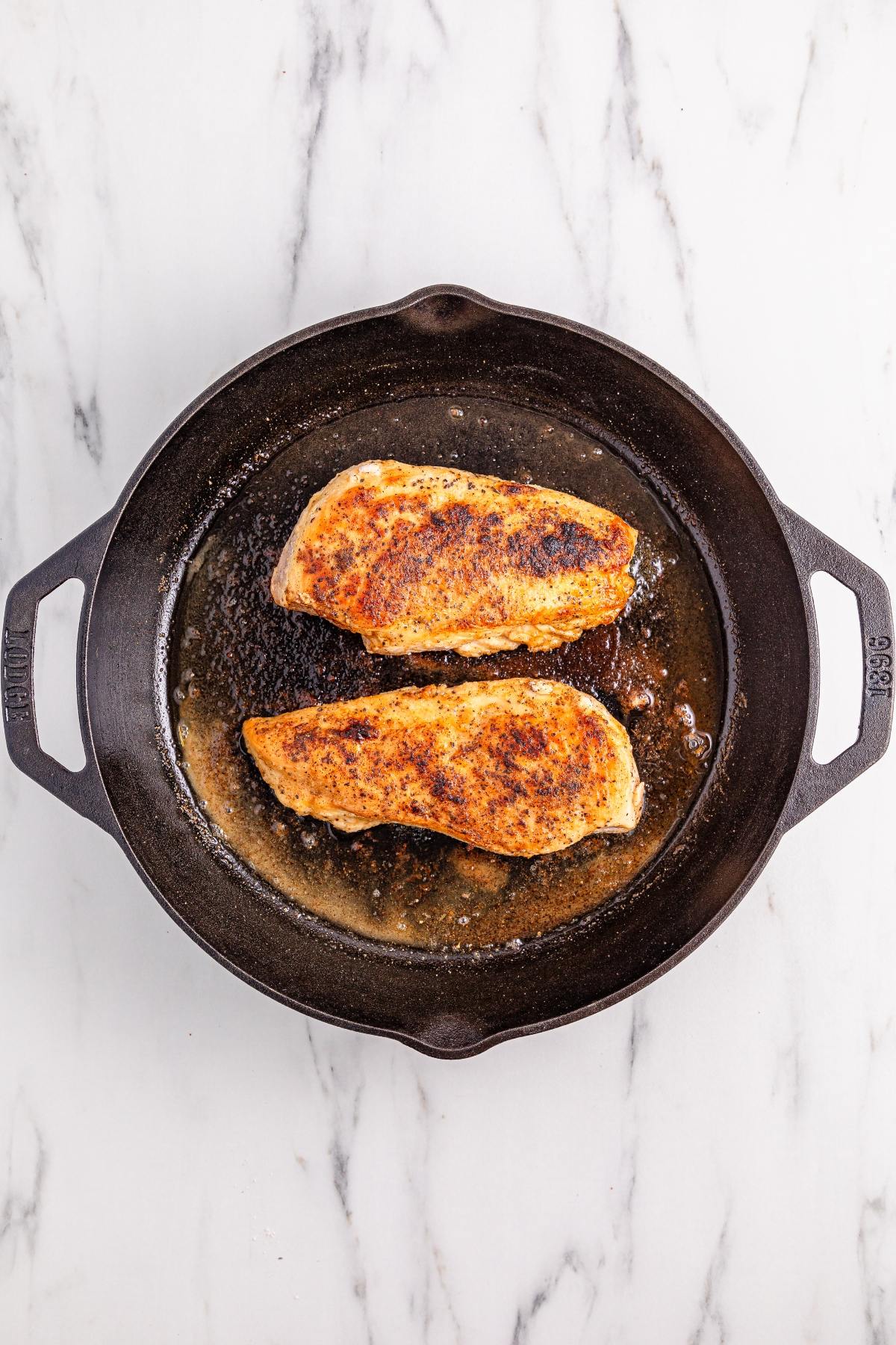 Two seasoned chicken breasts cooking in a black cast iron skillet on a white marble surface.