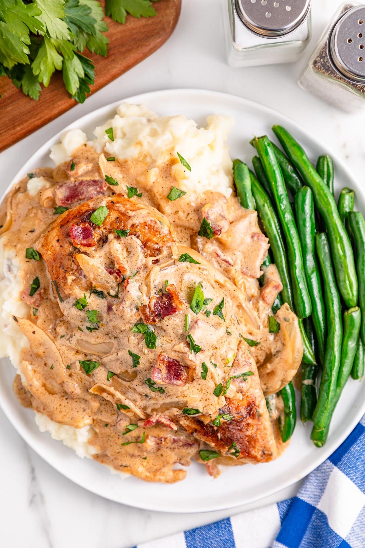 Plate of chicken with creamy sauce, mashed potatoes, and green beans, garnished with chopped parsley.