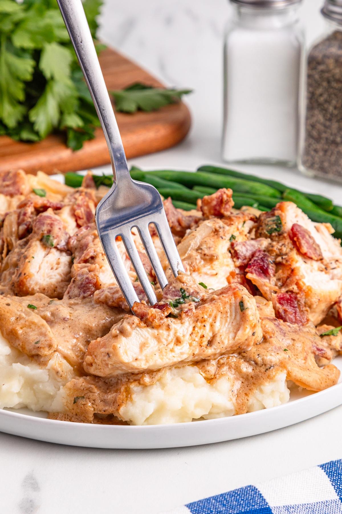 Fork cutting into creamy chicken with bacon on mashed potatoes, served with green beans on a white plate.