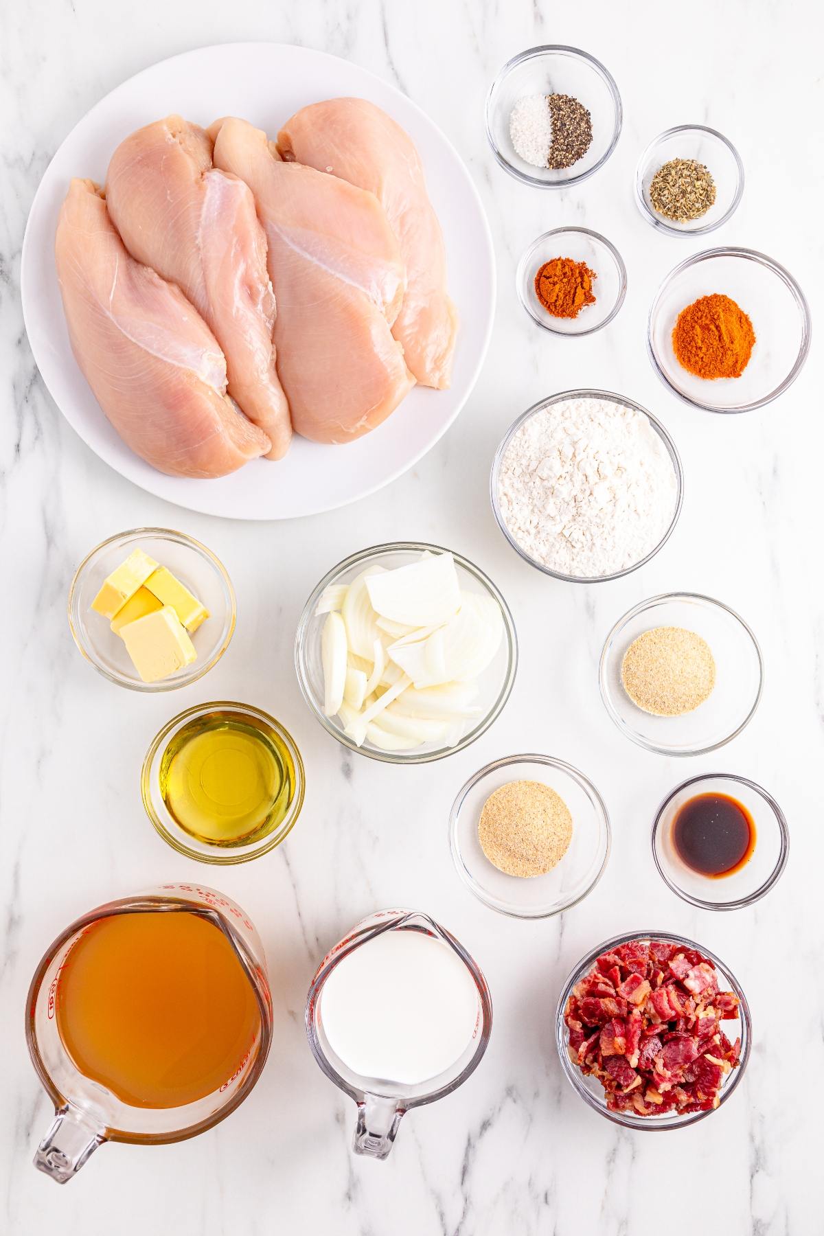 Raw chicken breasts, seasonings, sliced onions, butter, oil, flour, cream, broth, bacon bits, and sauces on a counter.