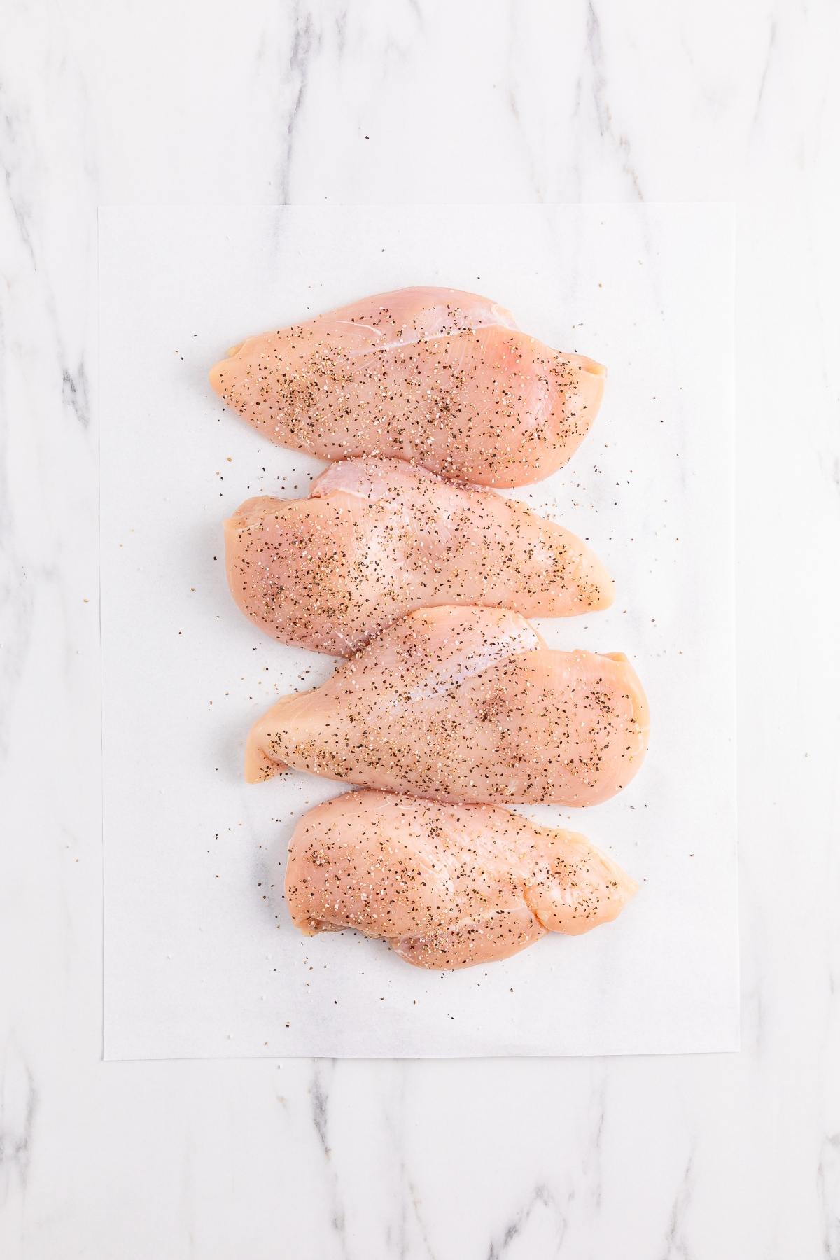 Four raw chicken breasts sprinkled with black pepper, arranged on white parchment paper on a marble surface.