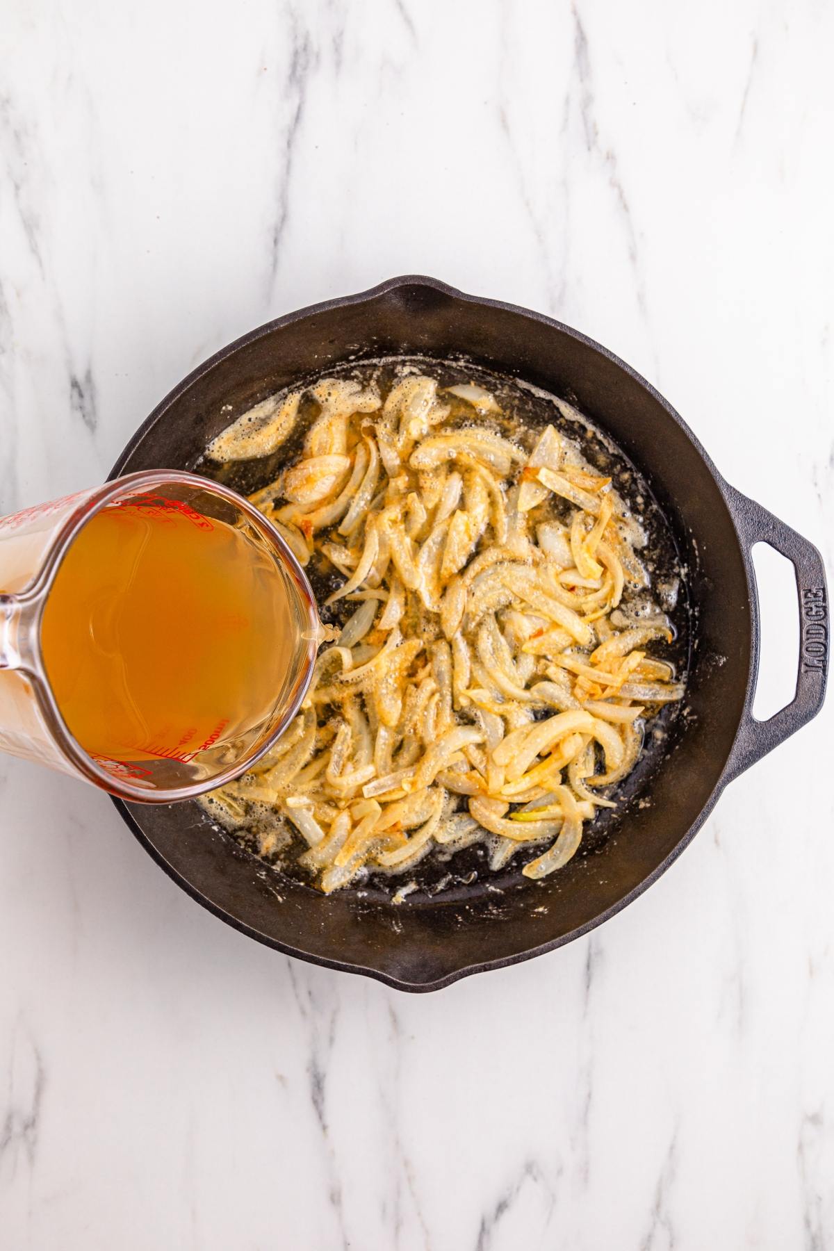 A glass measuring cup pours broth into a cast iron skillet with sautéed onions on a marble countertop.
