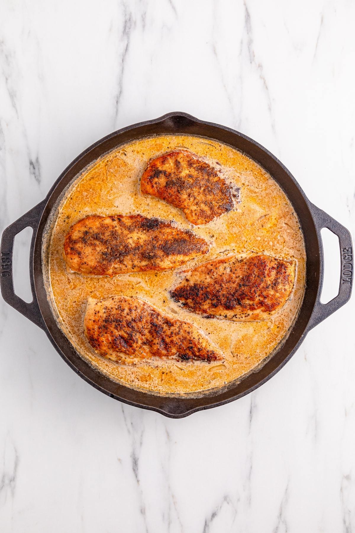 Four seasoned chicken breasts simmer in a creamy sauce in a round cast iron skillet on a marble surface.
