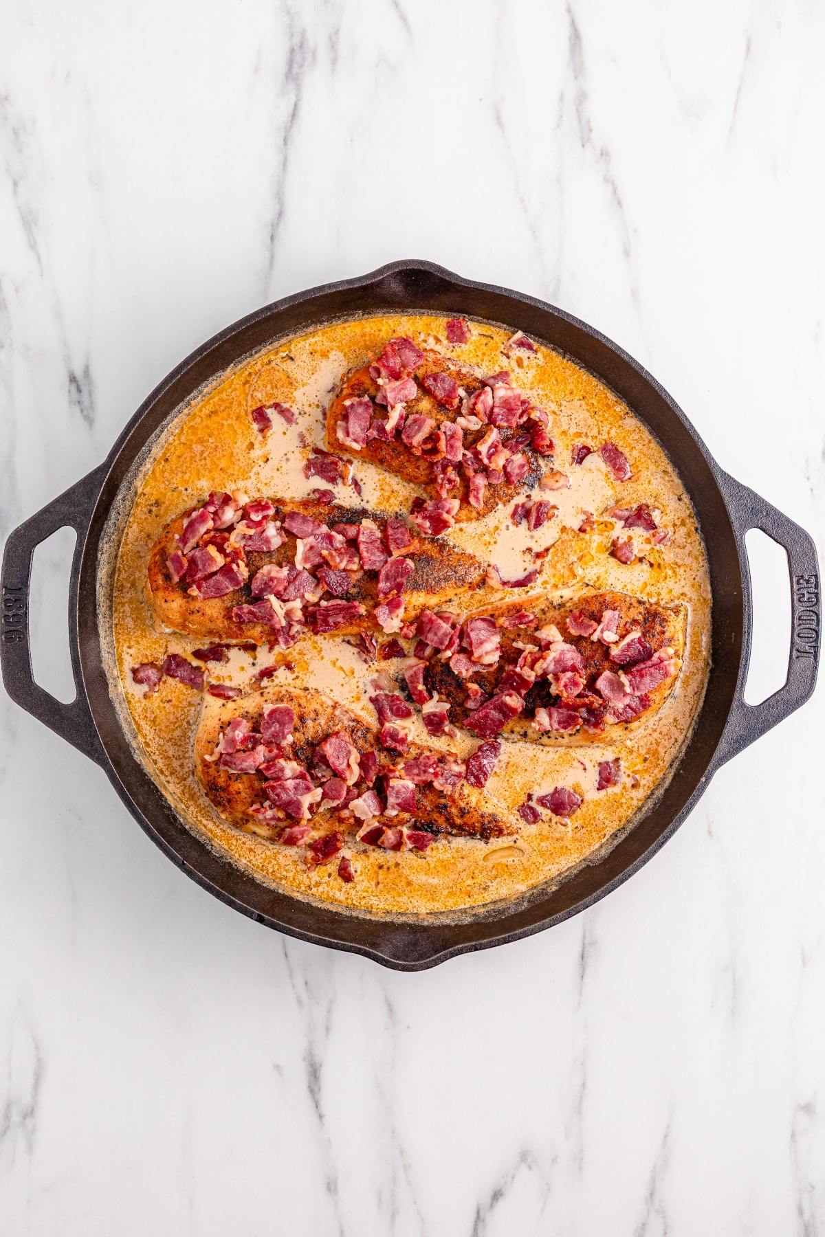 Cast iron skillet with creamy sauce and cooked chicken breasts topped with crispy bacon pieces.