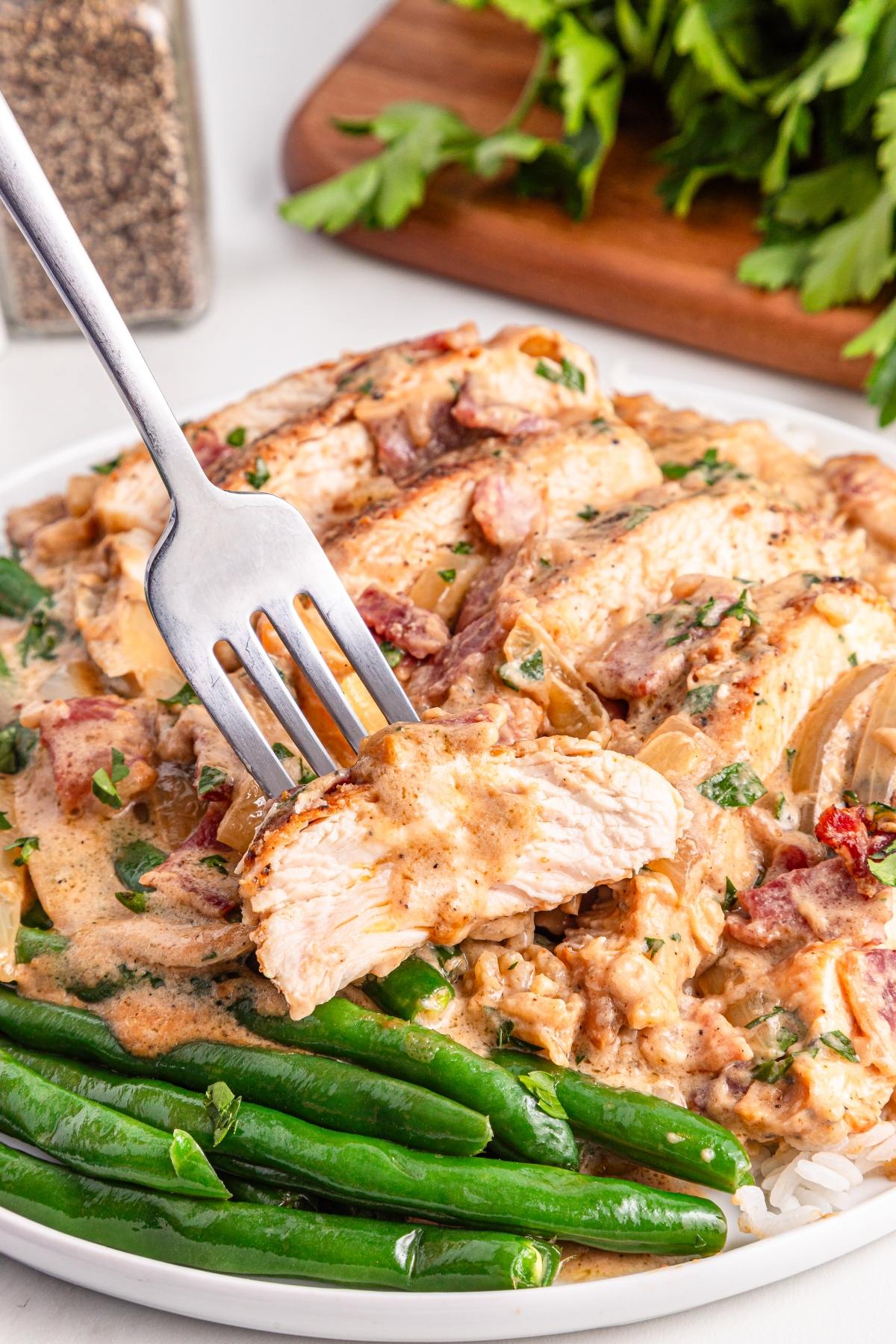 A fork holds a piece of creamy chicken over a plate with green beans and saucy chicken breast.