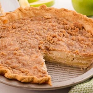 A sour cream apple pie with a crumb topping, baked in a pie tin with a slice removed, sits on a white surface.