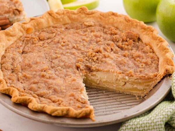 A sour cream apple pie with a crumb topping, baked in a pie tin with a slice removed, sits on a white surface.