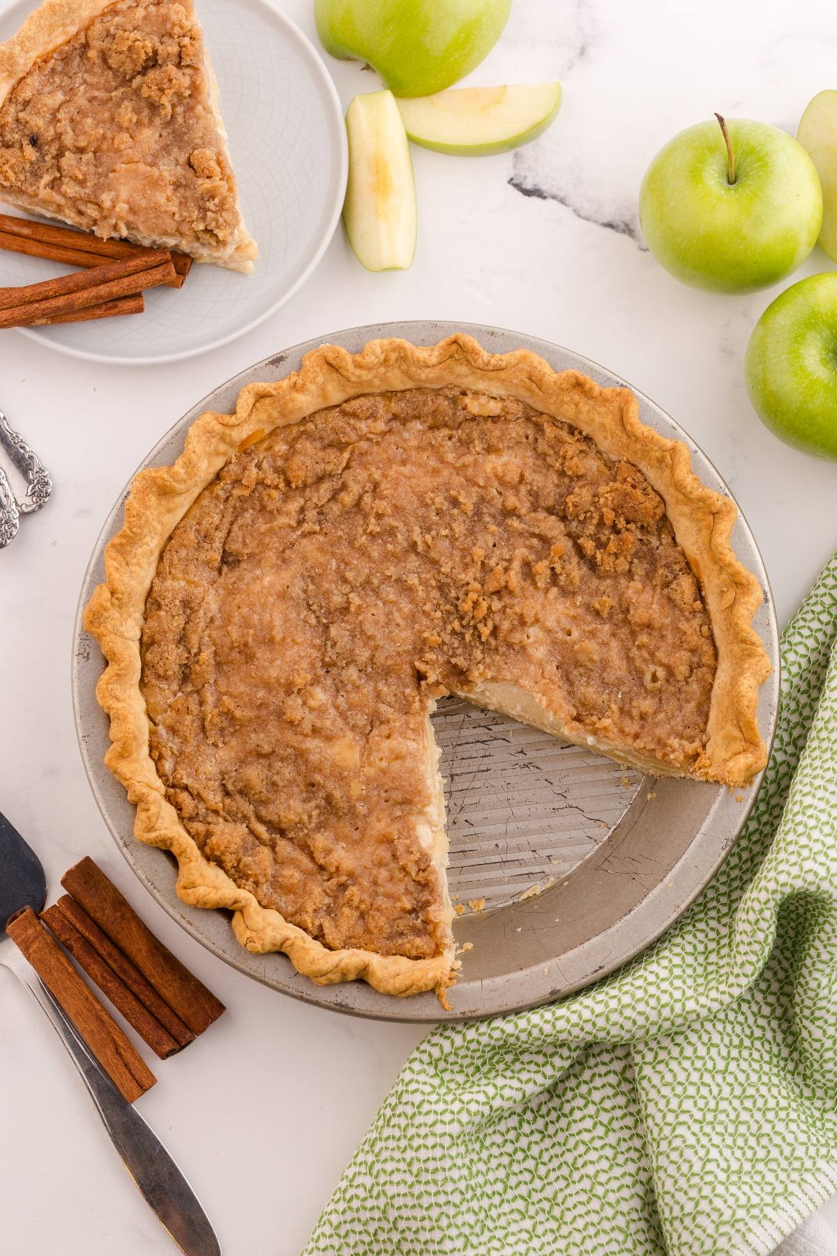 Whole apple pie with a crumbly topping, one slice missing, surrounded by green apples and cinnamon sticks.