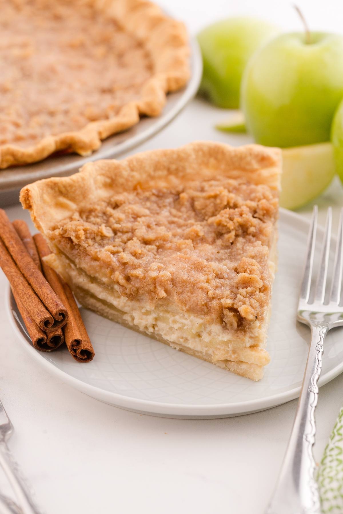 A slice of crumb-topped pie on a plate with cinnamon sticks and green apples in the background.