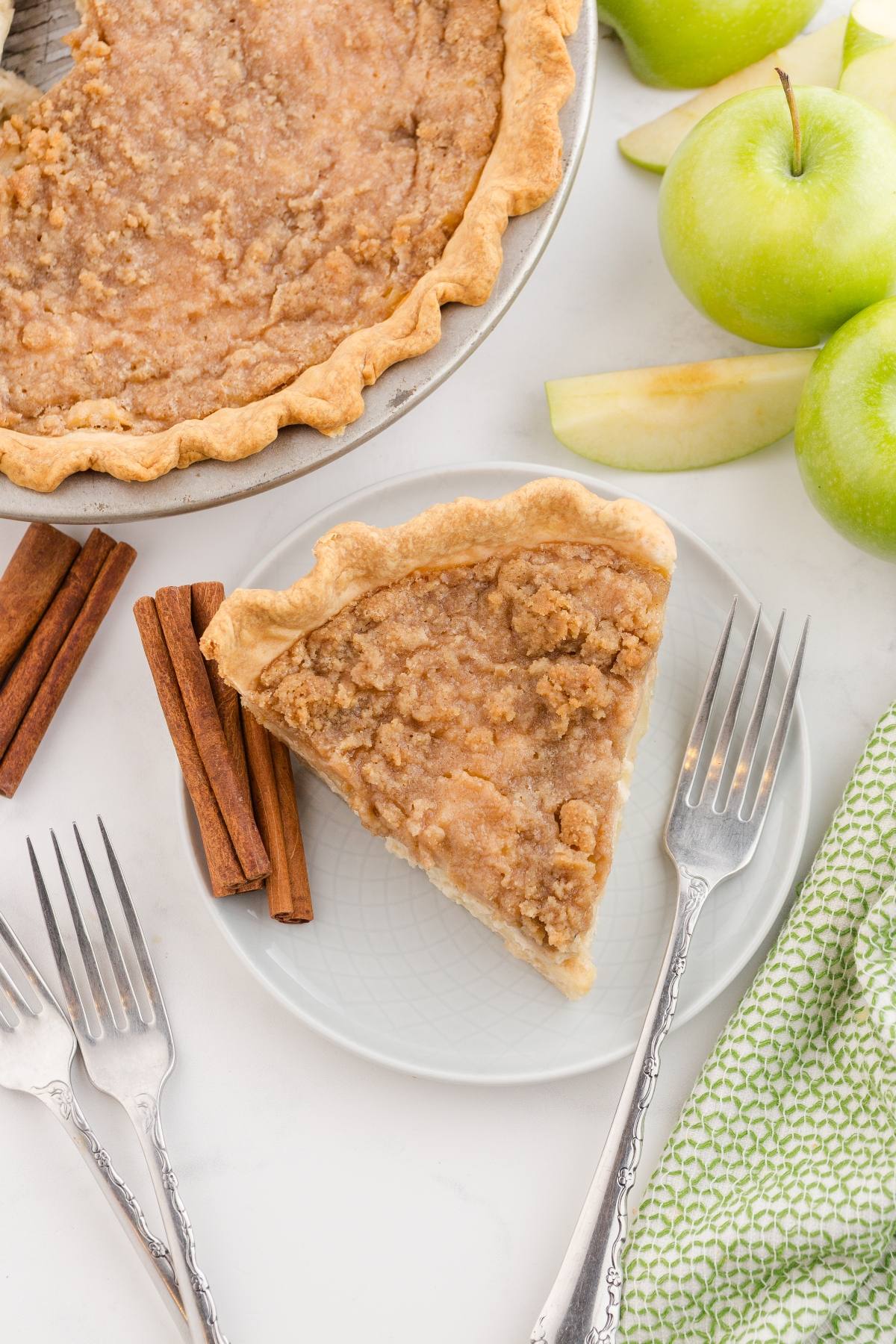 A slice of crumb-topped pie on a plate with forks, cinnamon sticks, and green apples nearby.