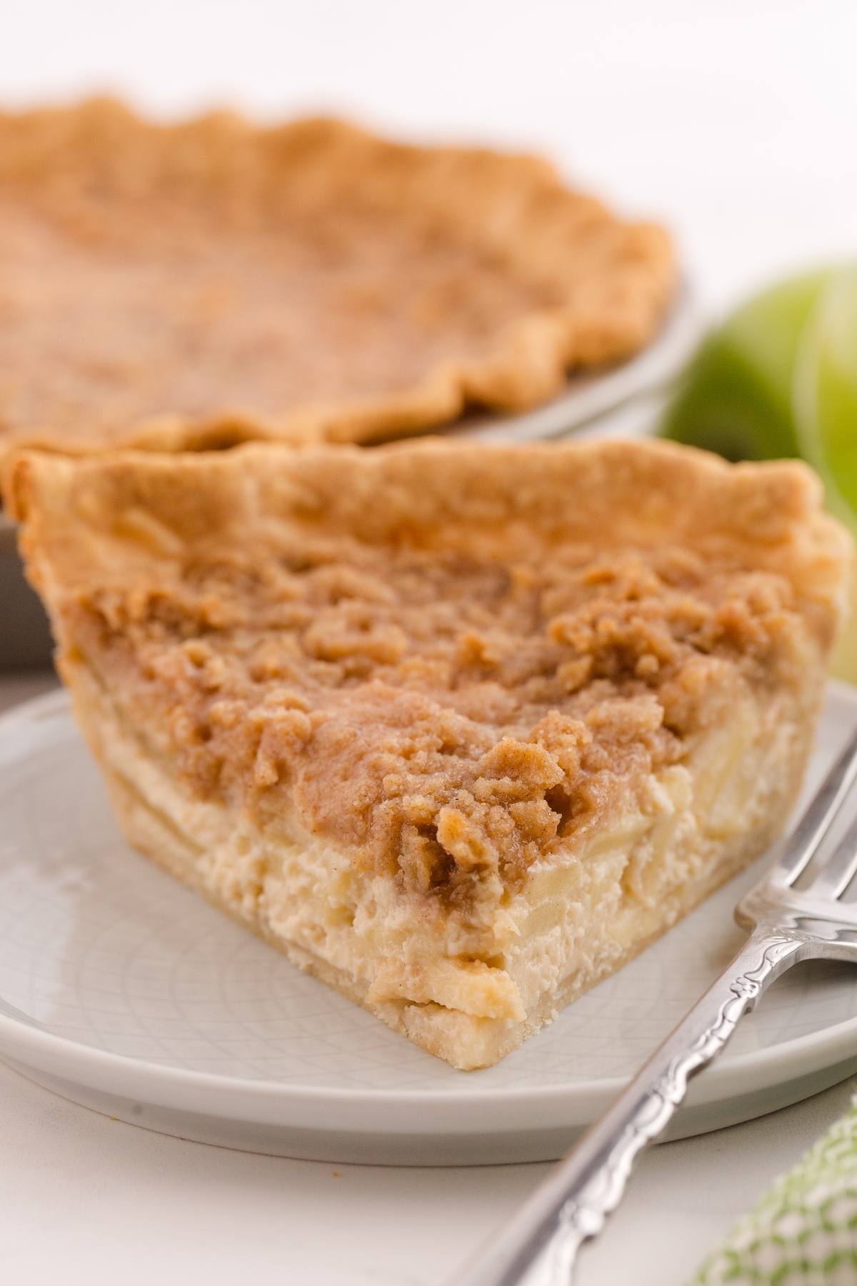 A slice of crumb-topped pie on a plate with a fork, with the whole pie and green apples in the background.