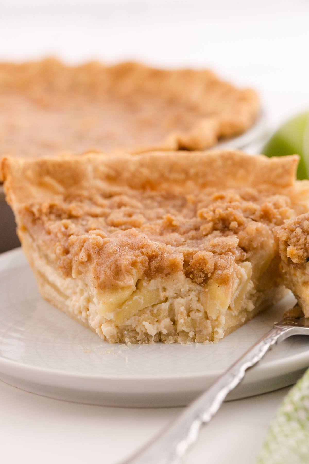 A slice of crumb-topped pie with apple filling on a white plate, with a pie in the background.