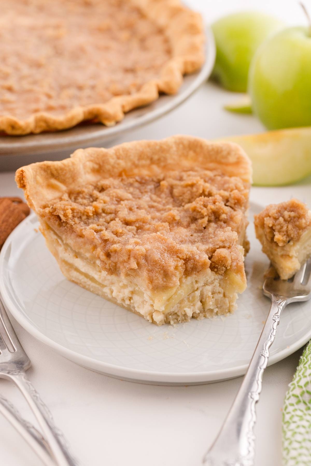 A slice of crumb-topped apple pie on a plate with a fork, with apples and more pie in the background.