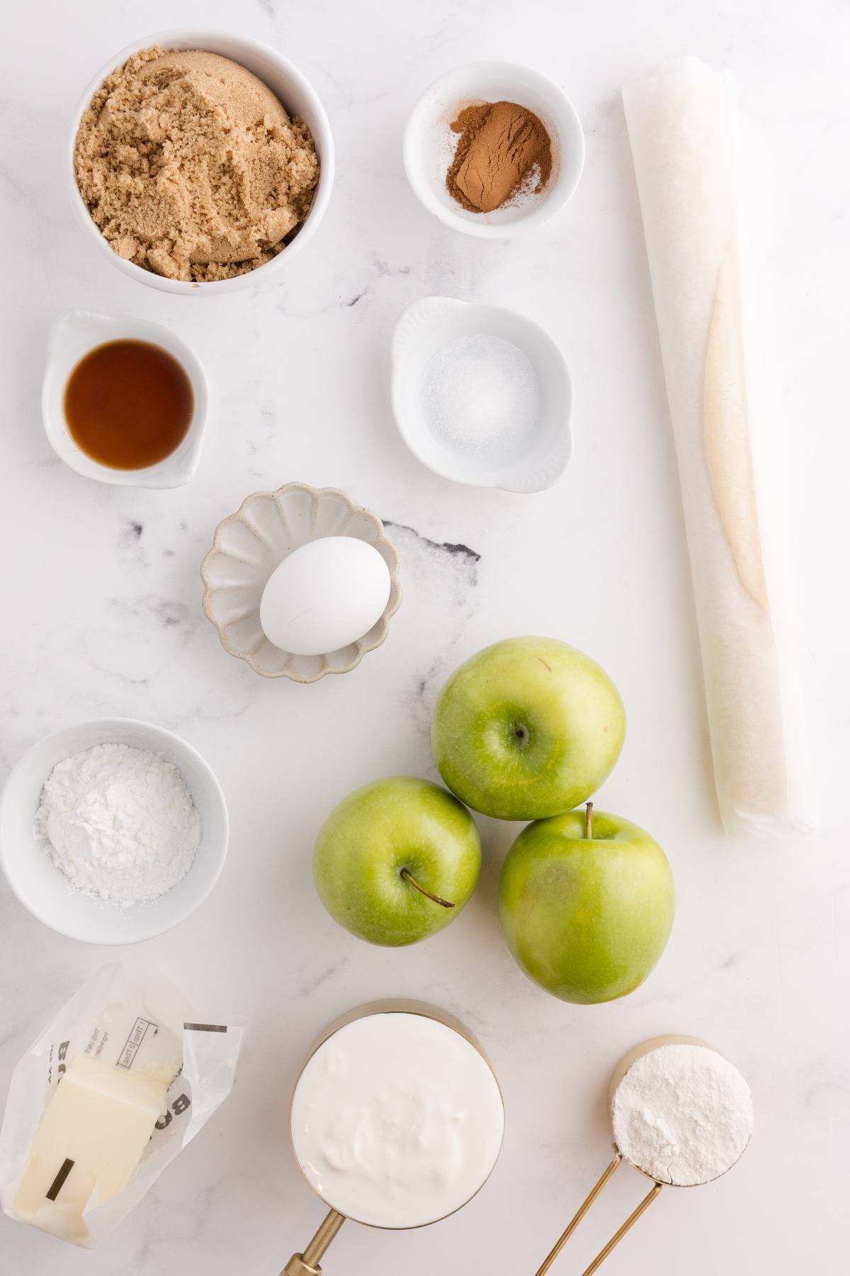 Baking ingredients on a white surface: green apples, egg, brown sugar, flour, butter, vanilla, cinnamon, and pie crust.