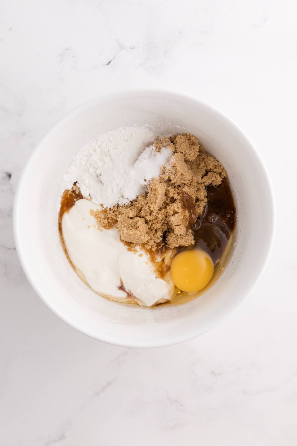 A white bowl with flour, brown sugar, sour cream, an egg, and vanilla on a white marble surface.