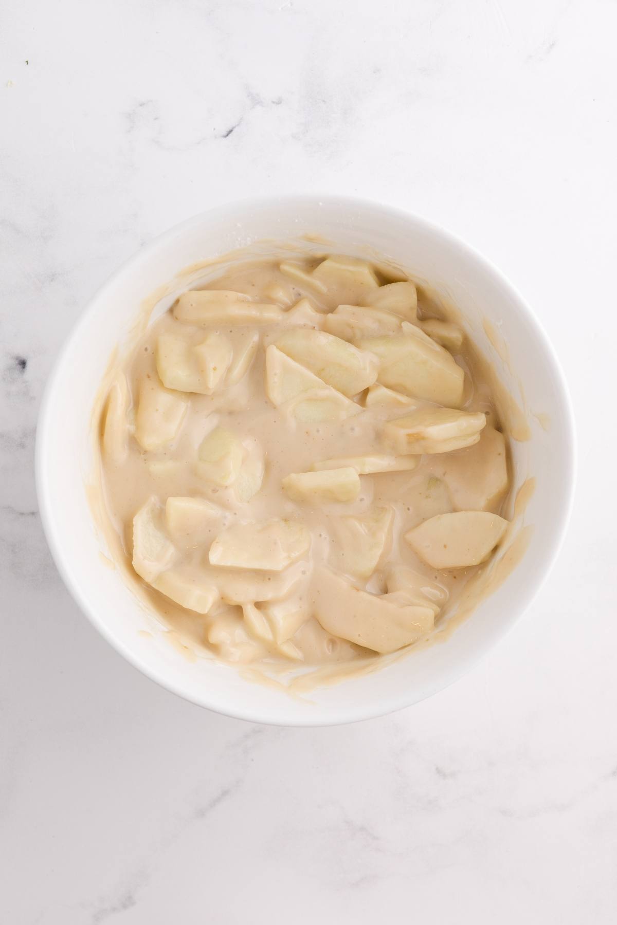 Sliced apples coated in a creamy, beige mixture inside a white bowl on a white marble surface.
