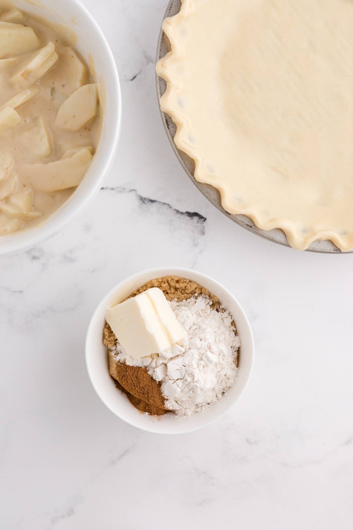 Bowl of pie filling, unbaked pie crust, and small bowl with butter, flour, sugar, and cinnamon on a white surface.