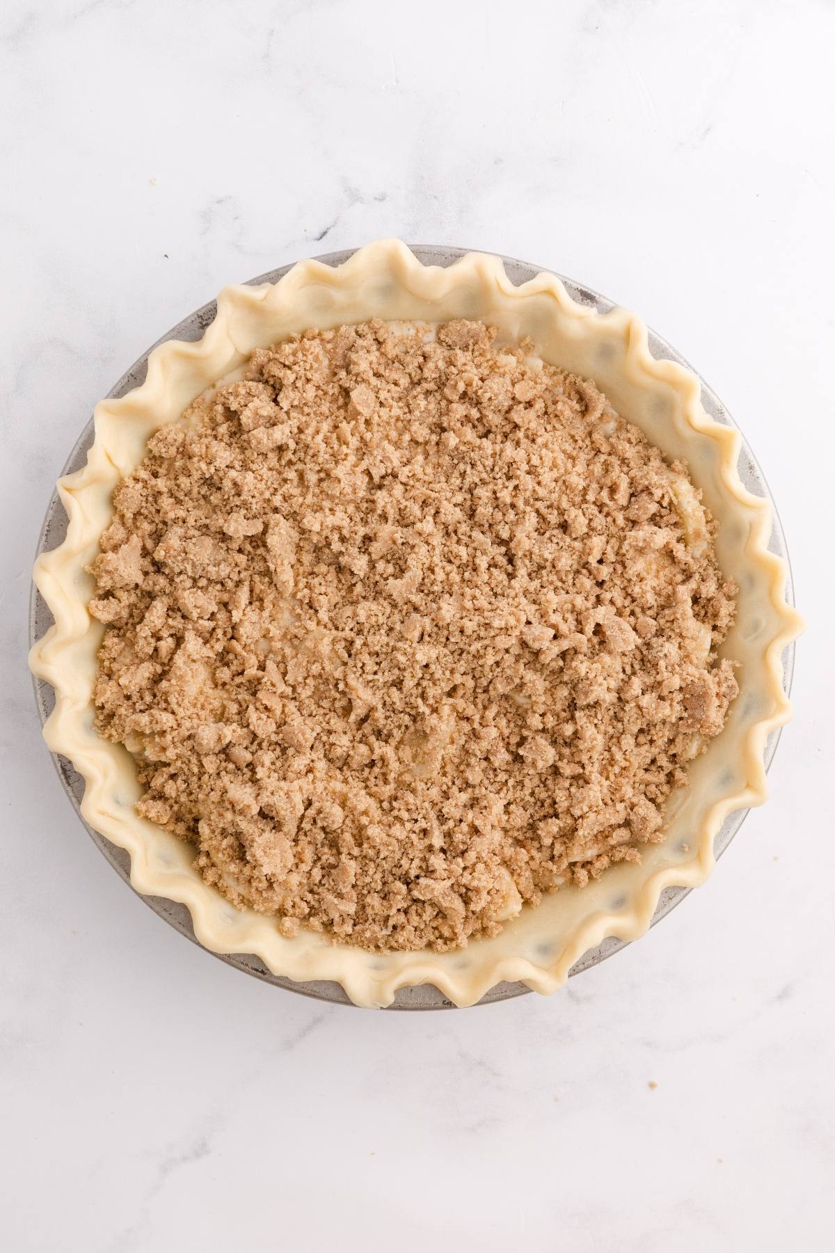 Unbaked pie with crimped edges and a crumbly streusel topping on a white marble surface.