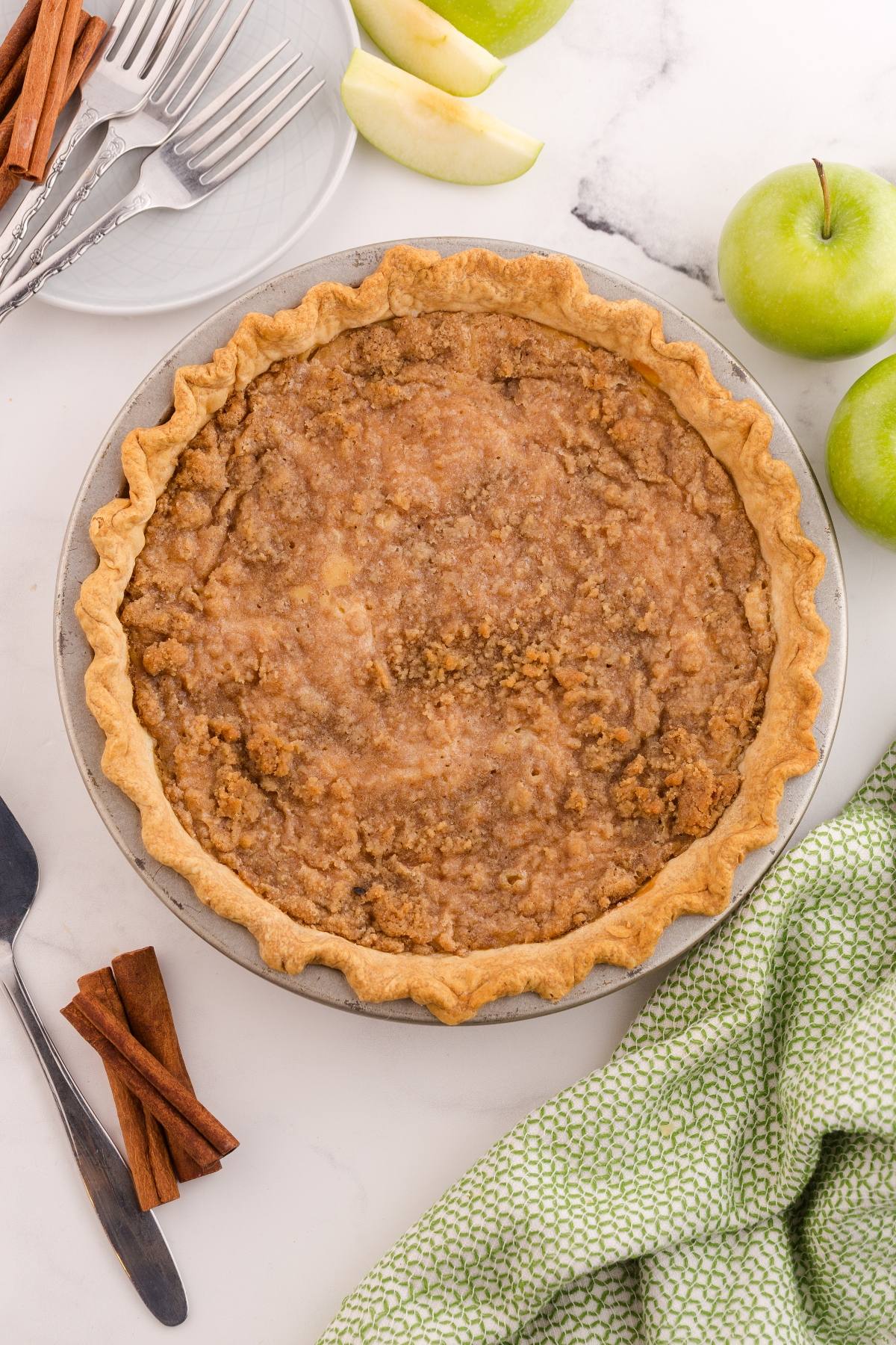 A baked apple pie with a crumbly topping, surrounded by green apples, cinnamon sticks, and utensils.