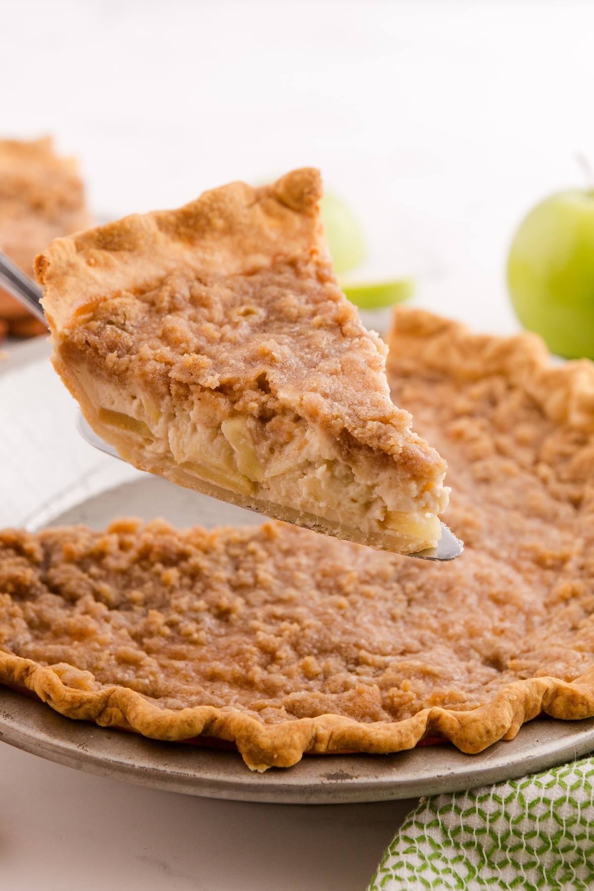 A slice of crumb-topped apple pie being lifted from a pie dish, with green apples in the background.