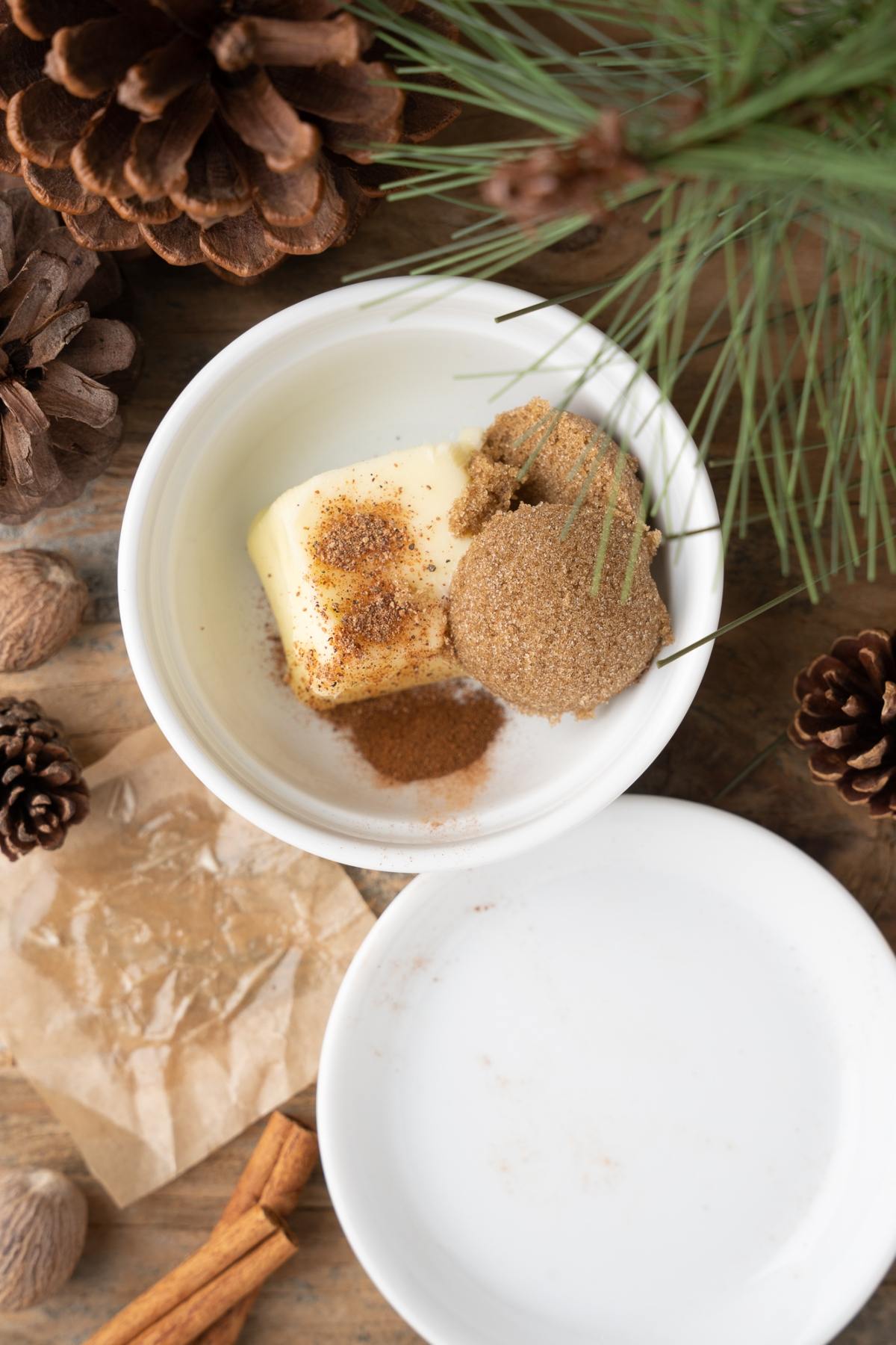 A bowl with butter, brown sugar, and spices sits on a wooden surface with pinecones and pine needles nearby—perfect ingredients for a cozy holiday cocktail or warm rum drink.