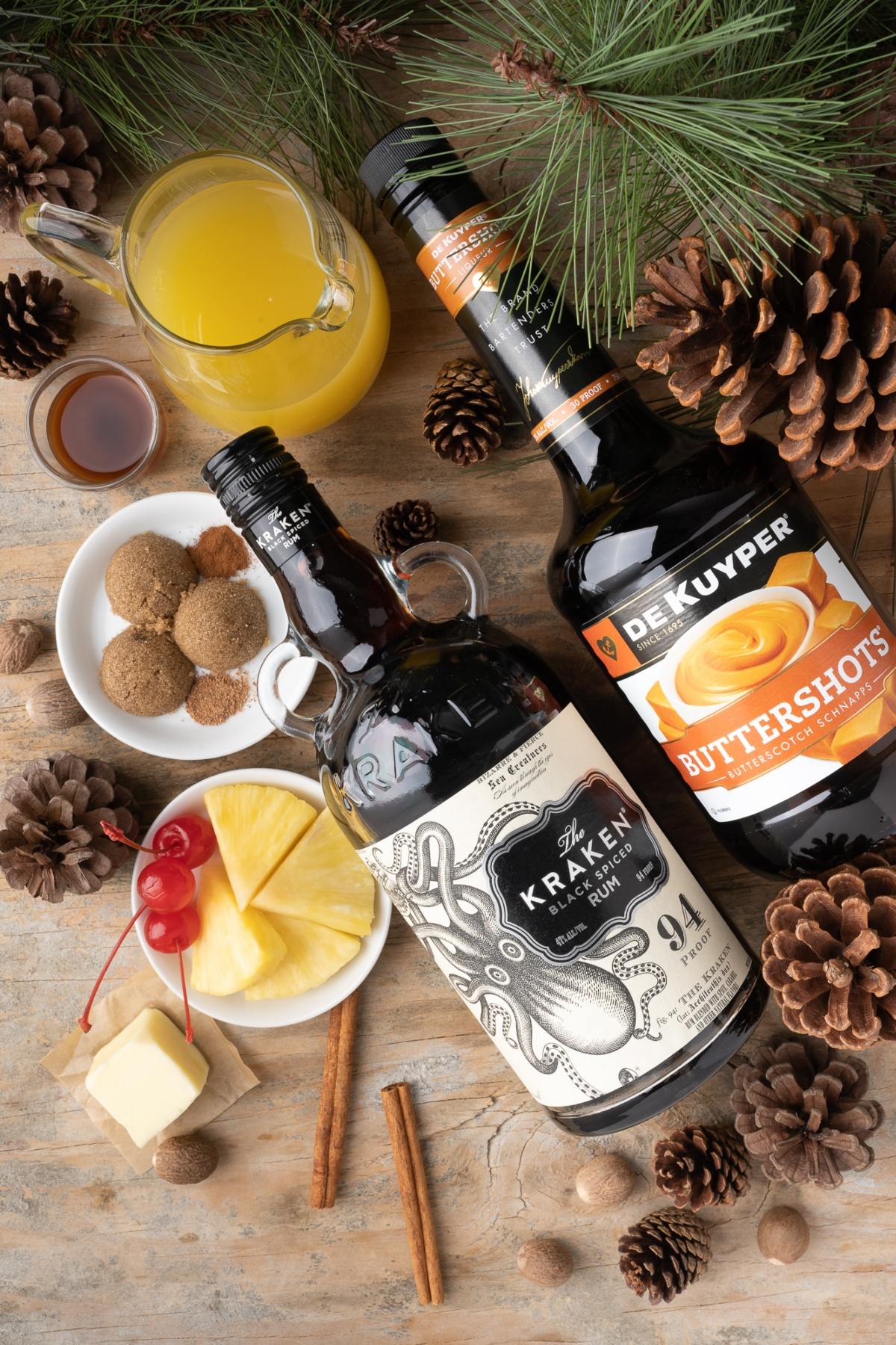 Two liquor bottles, fruit, spices, and a jug of juice arranged on a rustic table with pinecones and pine branches hint at a cozy Spiced Pineapple Hot Buttered Rum Toddy scene.