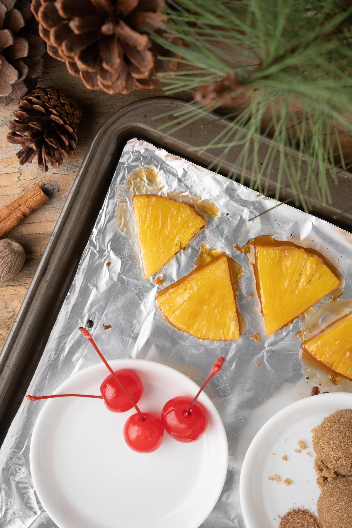 Four pineapple slices on foil, two maraschino cherries on a plate, and pinecones and pine needles nearby—perfect garnishes for a holiday cocktail like Spiced Pineapple Hot Buttered Rum Toddy with black spiced rum and butterscotch schnapps.
