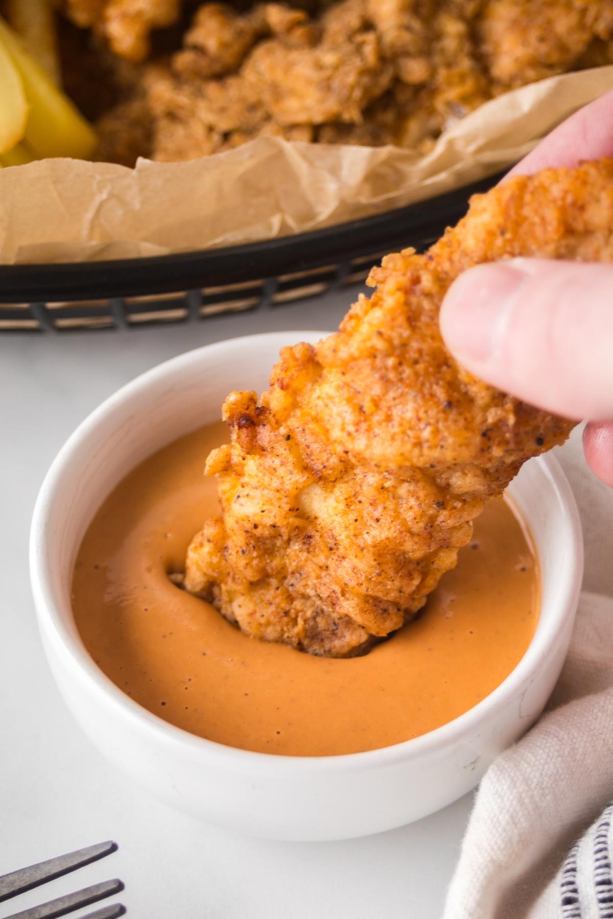 A hand dips a crispy chicken tender into a bowl of creamy dipping sauce.