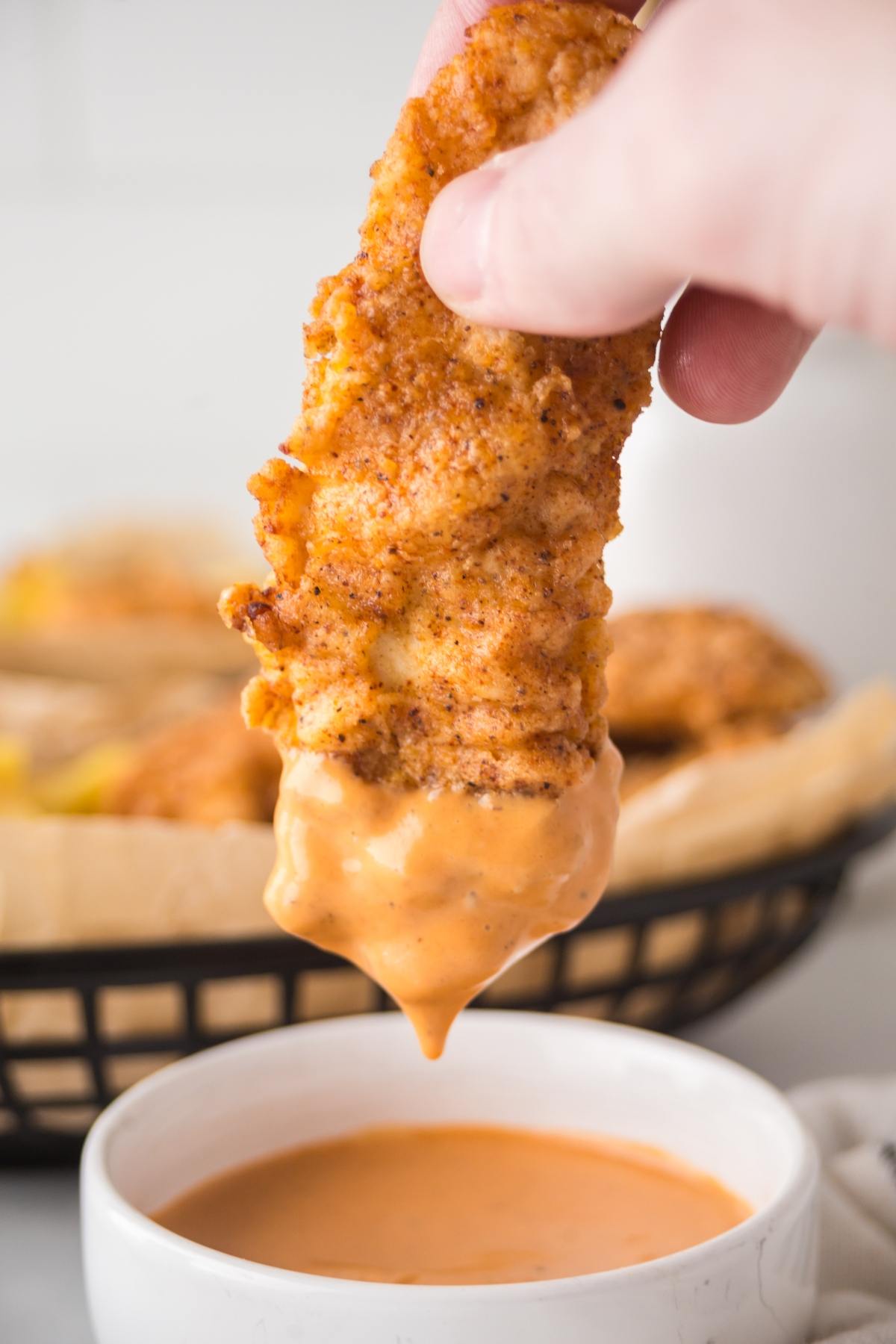 A hand dips a crispy chicken tender into a bowl of creamy orange dipping sauce.